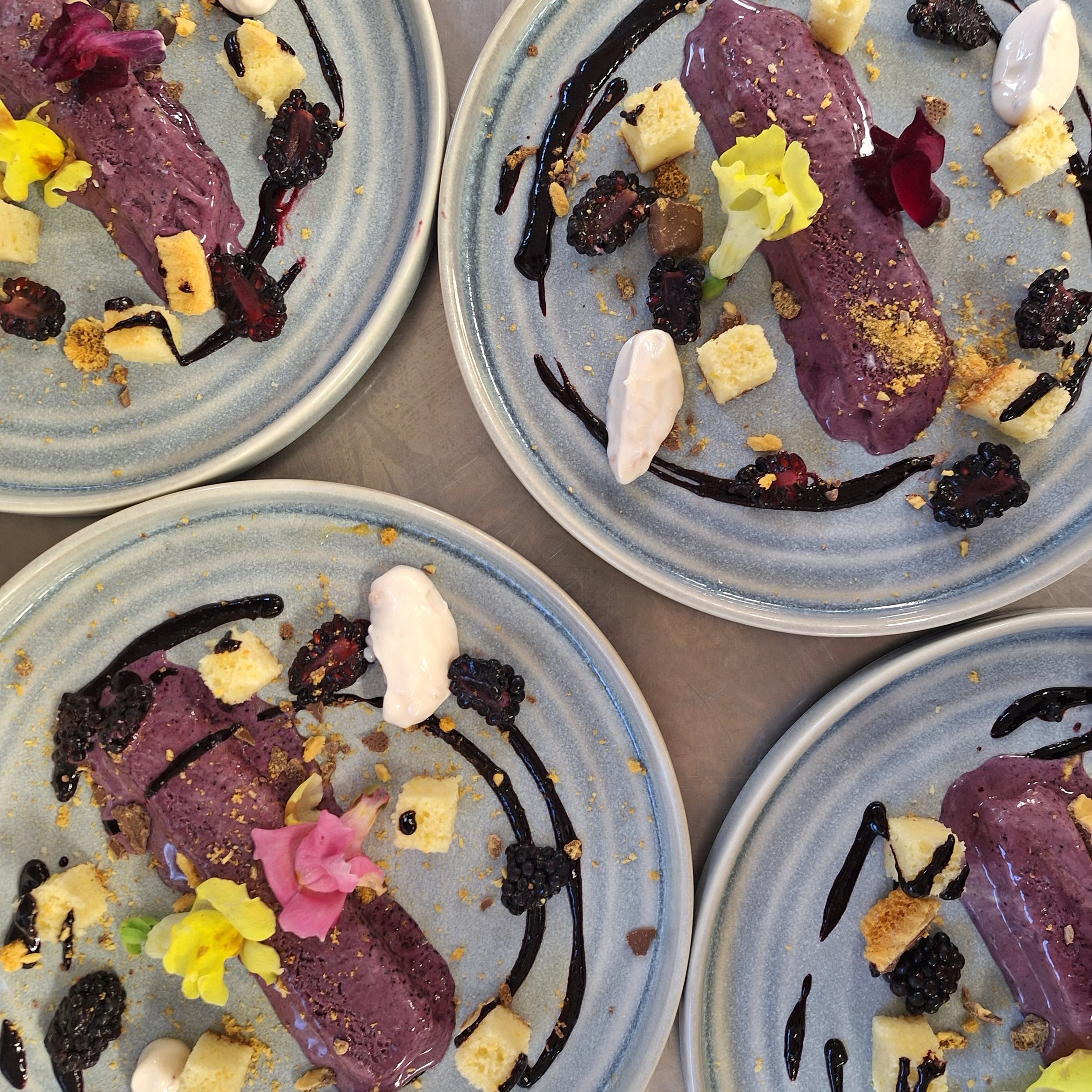 Dessert plates with purple ice cream, blackberries, white and pink edible flowers, crumbled cookies, and chocolate sauce