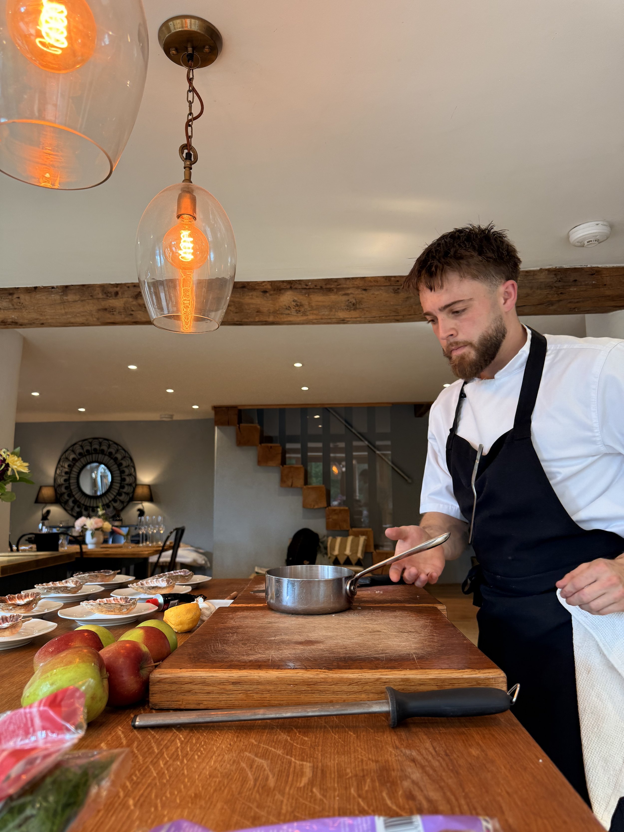 A chef with a beard and in a white shirt and black apron preparing food in a restaurant kitchen, standing at a wooden counter with apples, a saucepan, and a knife. The restaurant has wooden beams, decorative lighting, and a mirror on the wall.