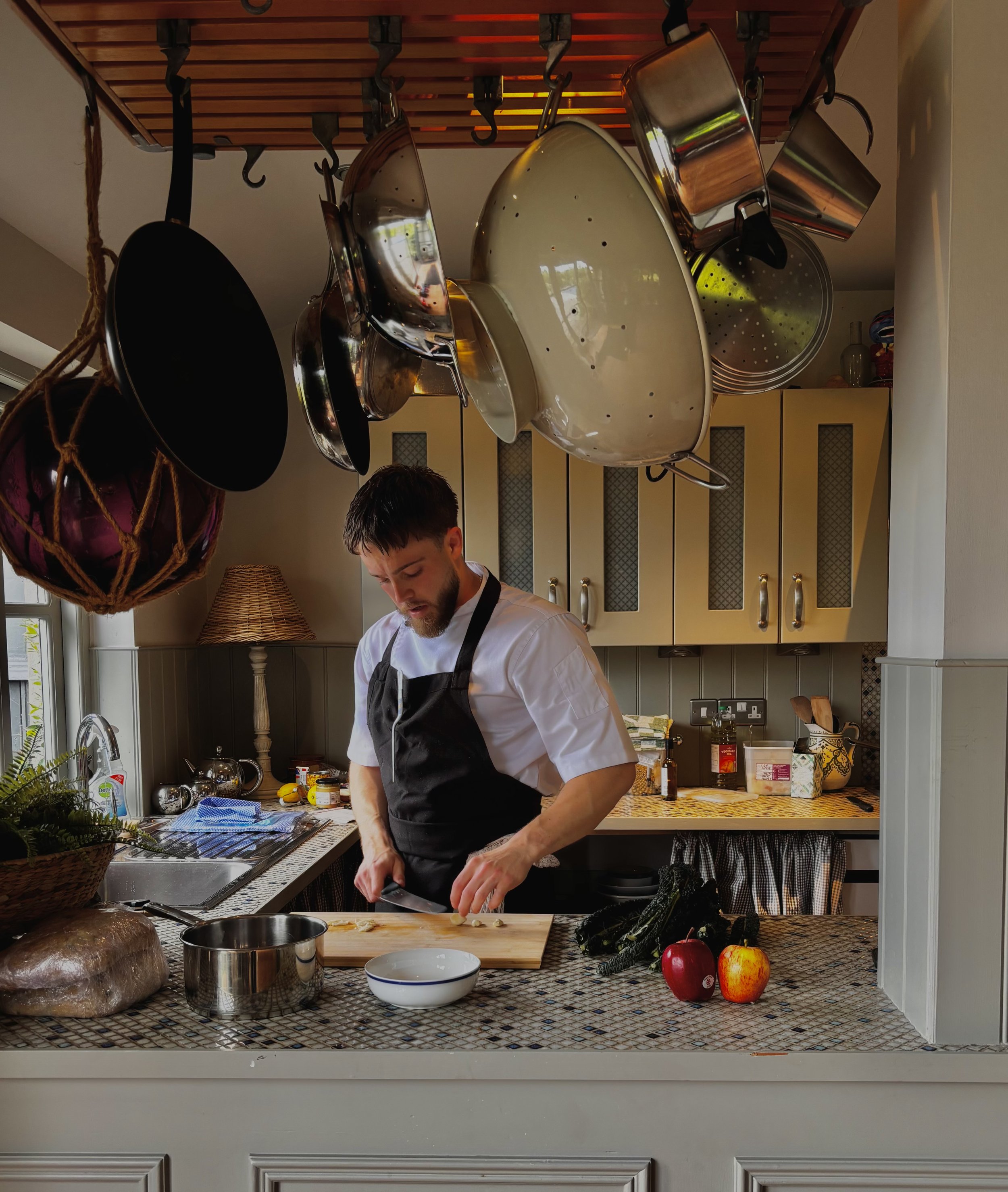 A man in a white shirt and black apron chopping vegetables at a kitchen counter. The kitchen has hanging pots and pans, a window with sunlight, and various kitchen items and fruits like peppers, apples, and a zucchini on the counter.