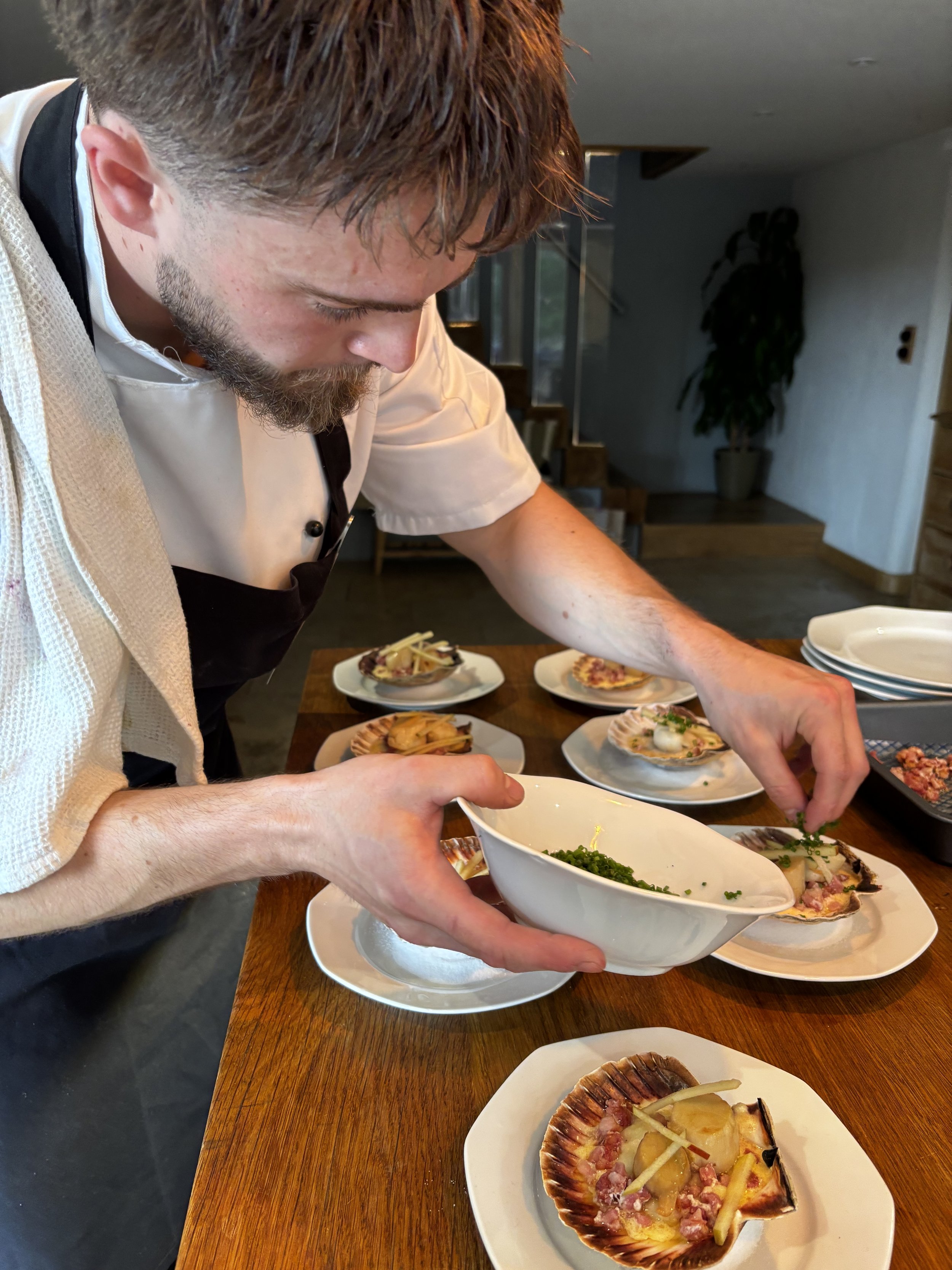 A chef garnishing plates of food in a kitchen. The table has multiple dishes, including a shell-shaped dish with toppings, and a bowl of chopped herbs or greens.