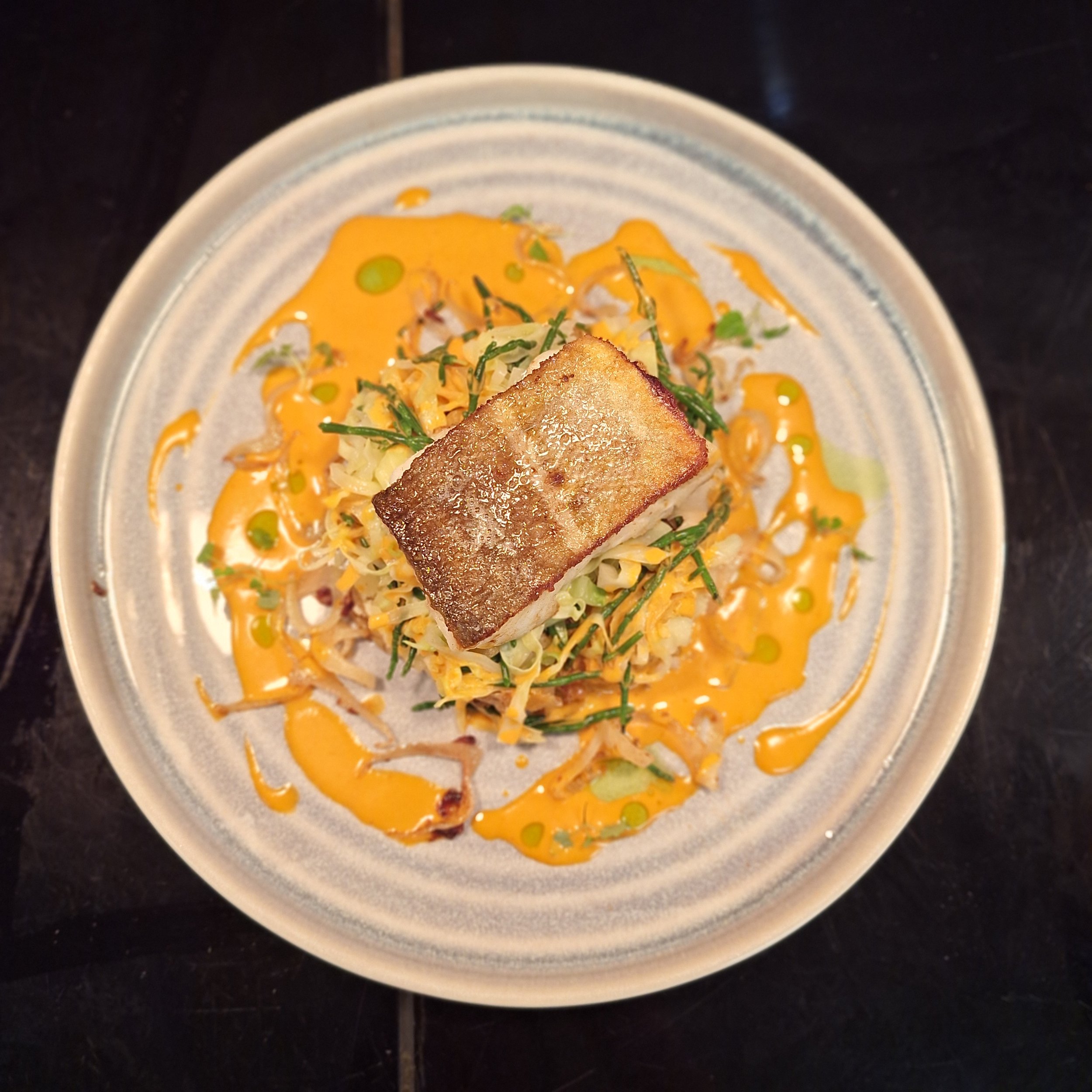 A plated serving of grilled fish with crispy skin, topped with sautéed vegetables and garnished with a drizzle of orange sauce and green herbs.