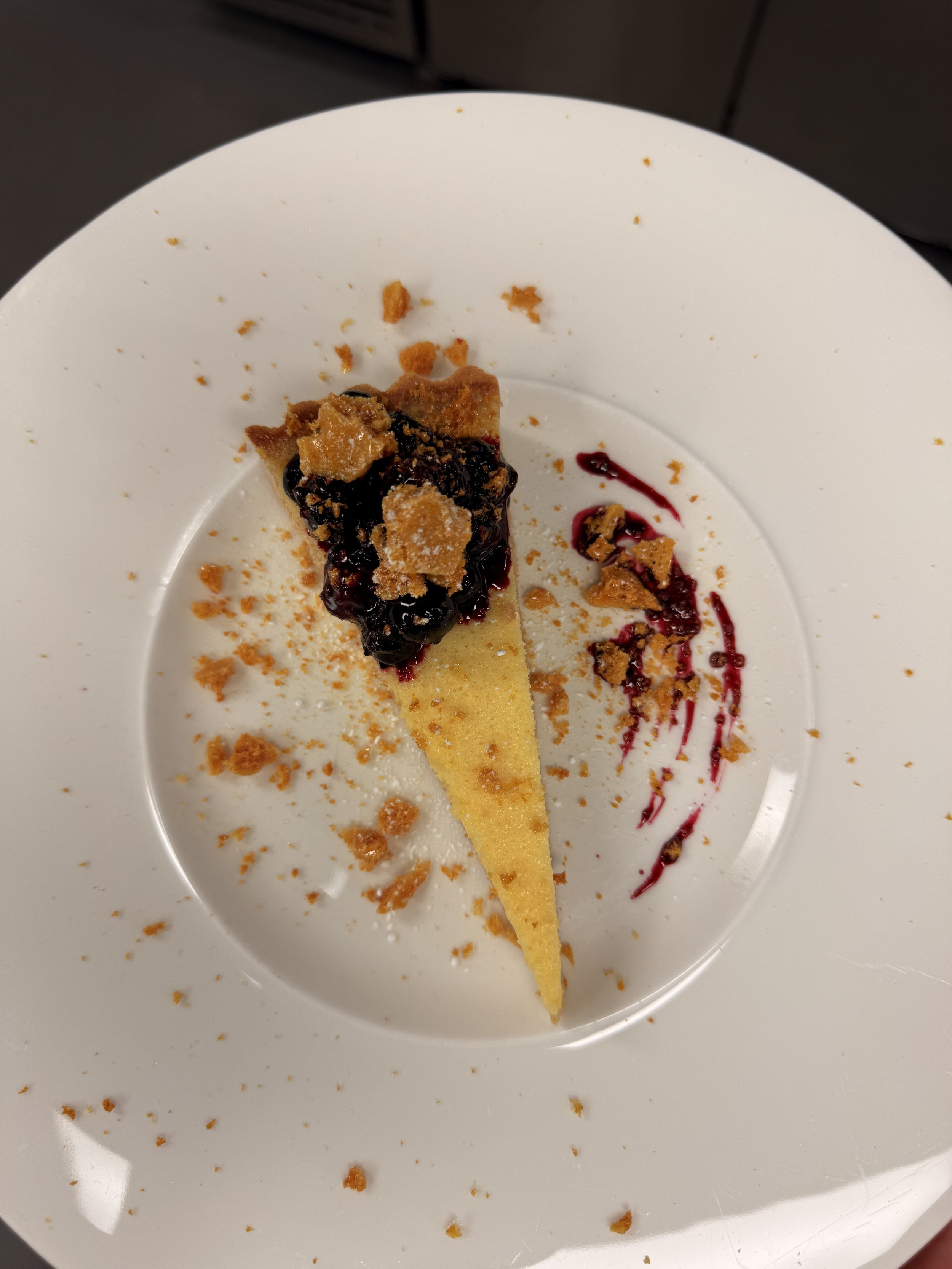 A slice of cheesecake topped with blueberry compote and crumbled cookies on a white plate with berry sauce and crumbs.