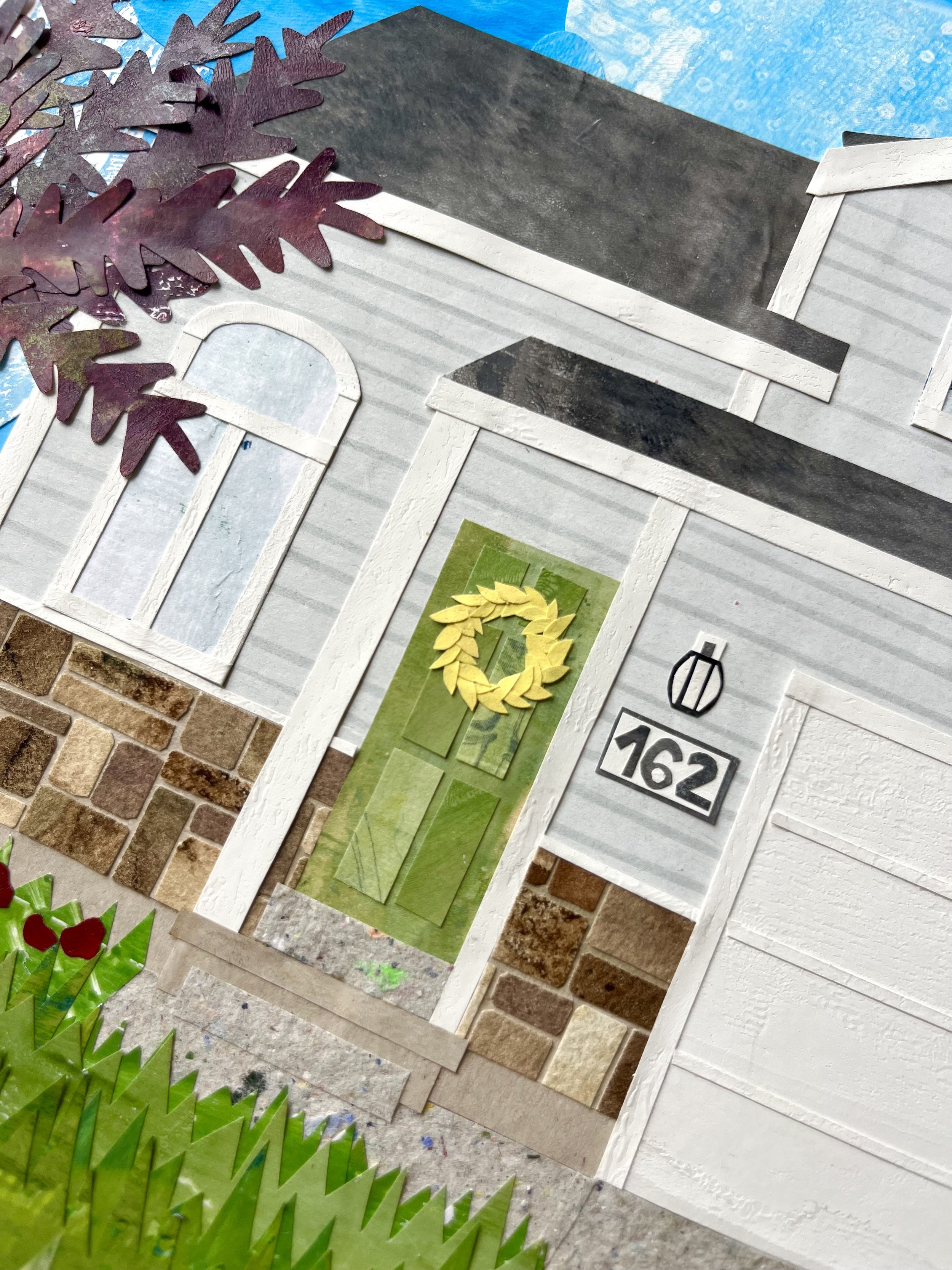 Close-up of a decorative house facade with a green door, brick and vinyl siding, and a yellow wreath. There is a house number 162, an outdoor light fixture, a window, and purple tree leaves at the top left. The image appears to be part of a scrapbook or collage with layered paper and textured materials.