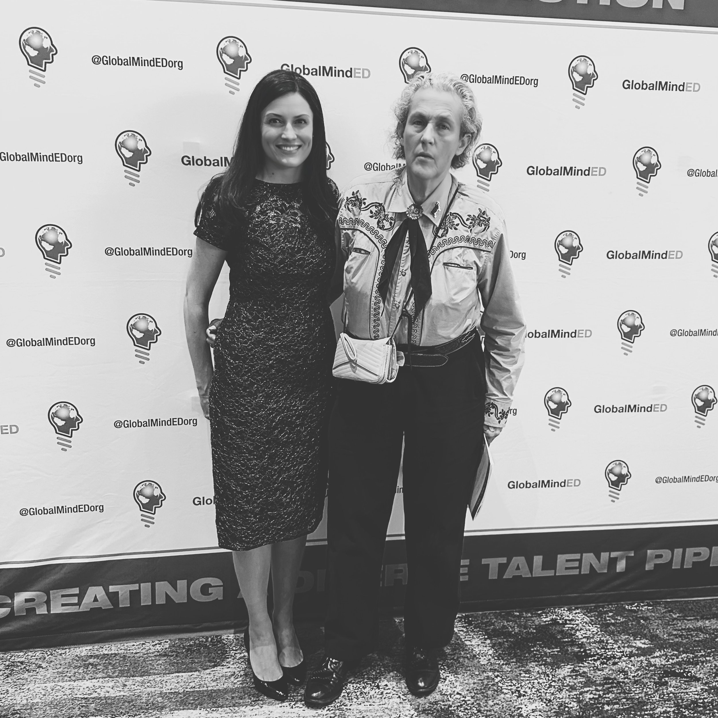 Two women standing side by side, smiling at an event with a backdrop displaying the words 'Global MindED' and a logo of a light bulb with a brain inside. One woman is wearing a patterned dress and high heels, the other is dressed in a Western-style shirt and dark pants.