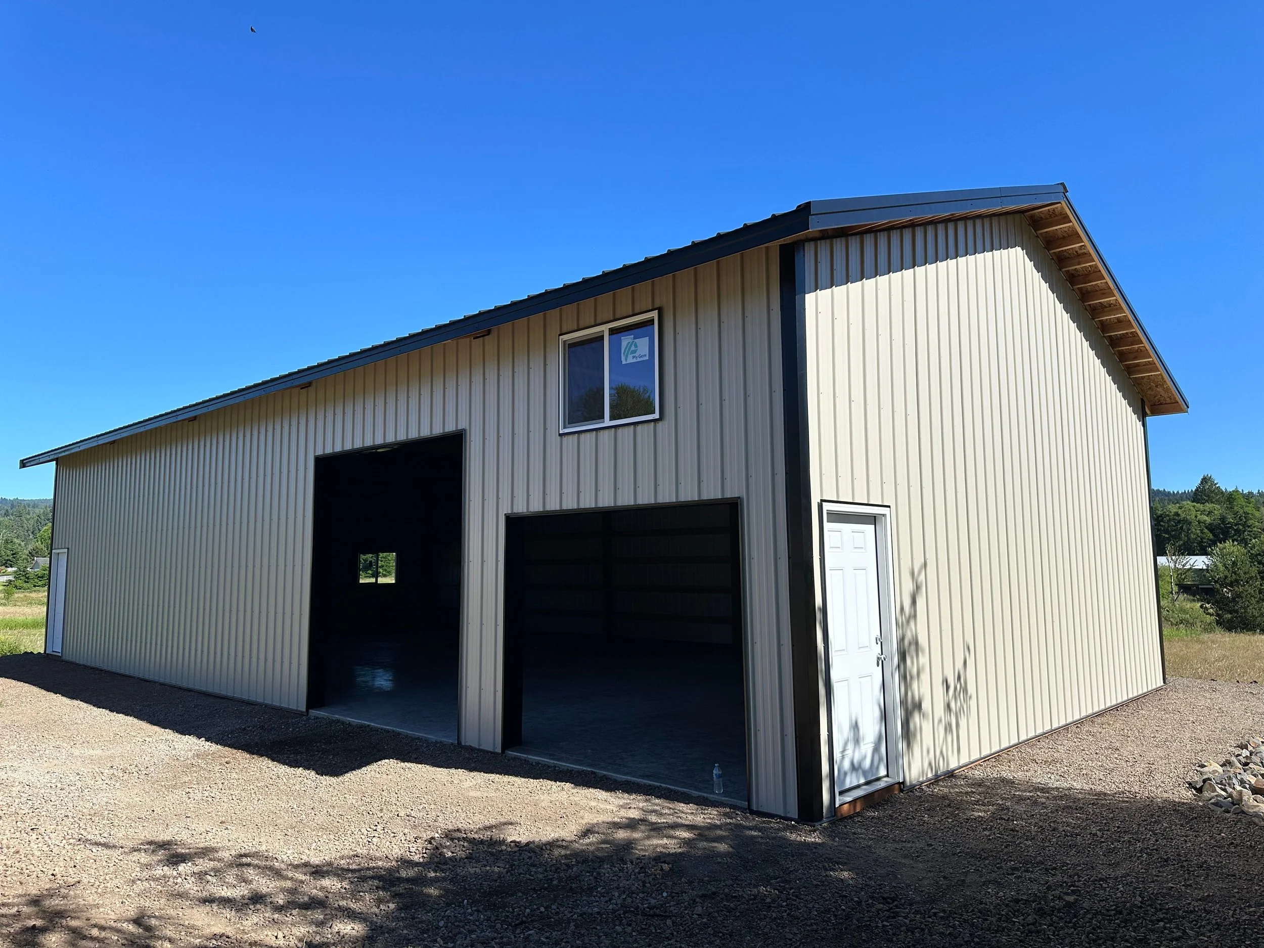 A large metal storage building with beige siding, black trim, a white door, and a small window, situated on a gravel lot under a clear blue sky.