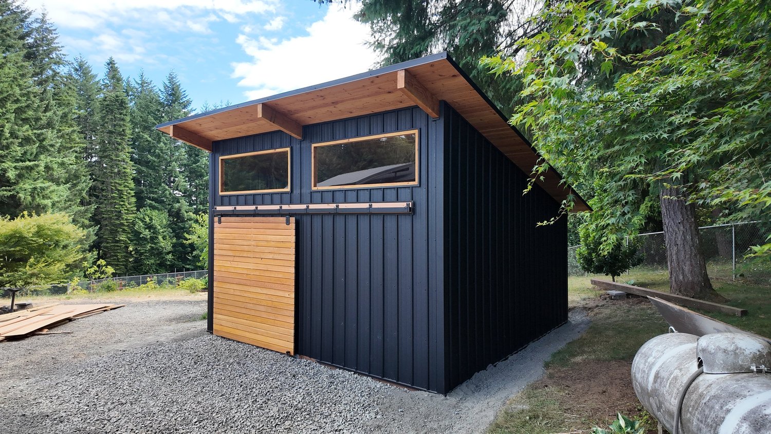 Modern custom post-frame building with black vertical steel siding, a mono-slope roof, cedar sliding door, and clerestory windows in a wooded Southwest Washington lot