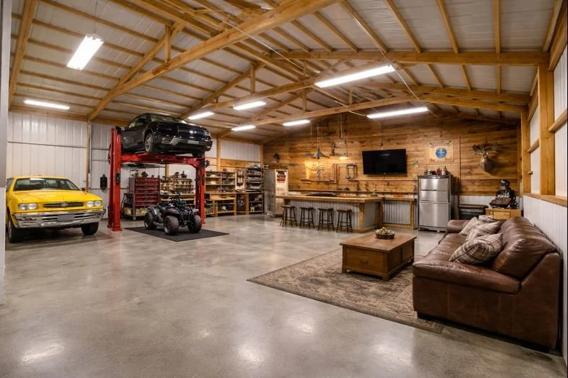 A spacious garage with a car lift holding a black sports car, a yellow classic car parked on the floor, a small black ATV, and a well-organized workshop area with tools and shelves. Adjacent to the workshop, there's a cozy living space with a leather sofa, coffee table, and a wall-mounted TV.