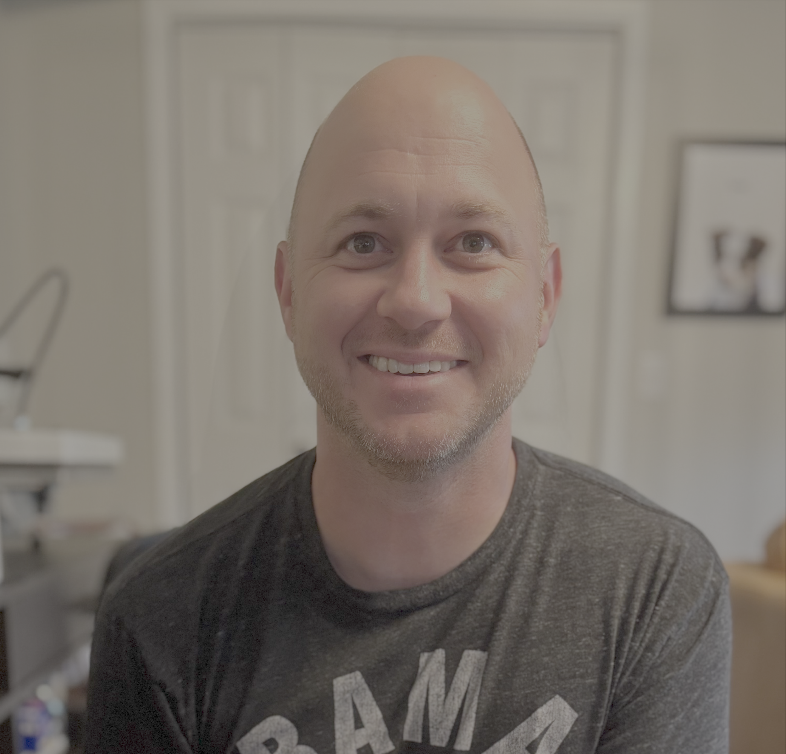 A smiling man with a bald head and light beard, wearing a dark gray t-shirt in a room with light-colored walls and a framed picture in the background.