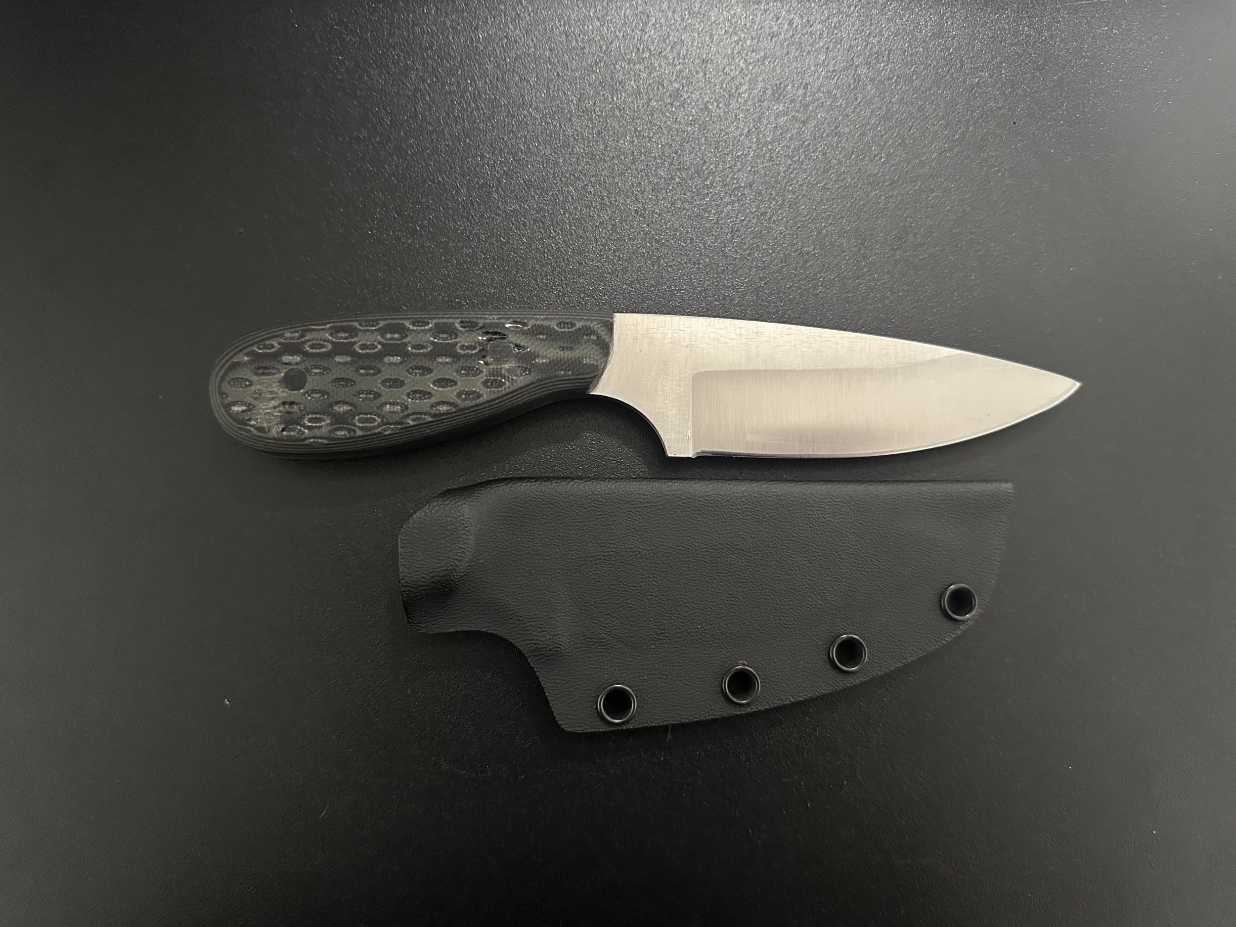 A small fixed-blade knife with a textured black handle and a stainless steel blade, resting on a black surface with a black sheath underneath.