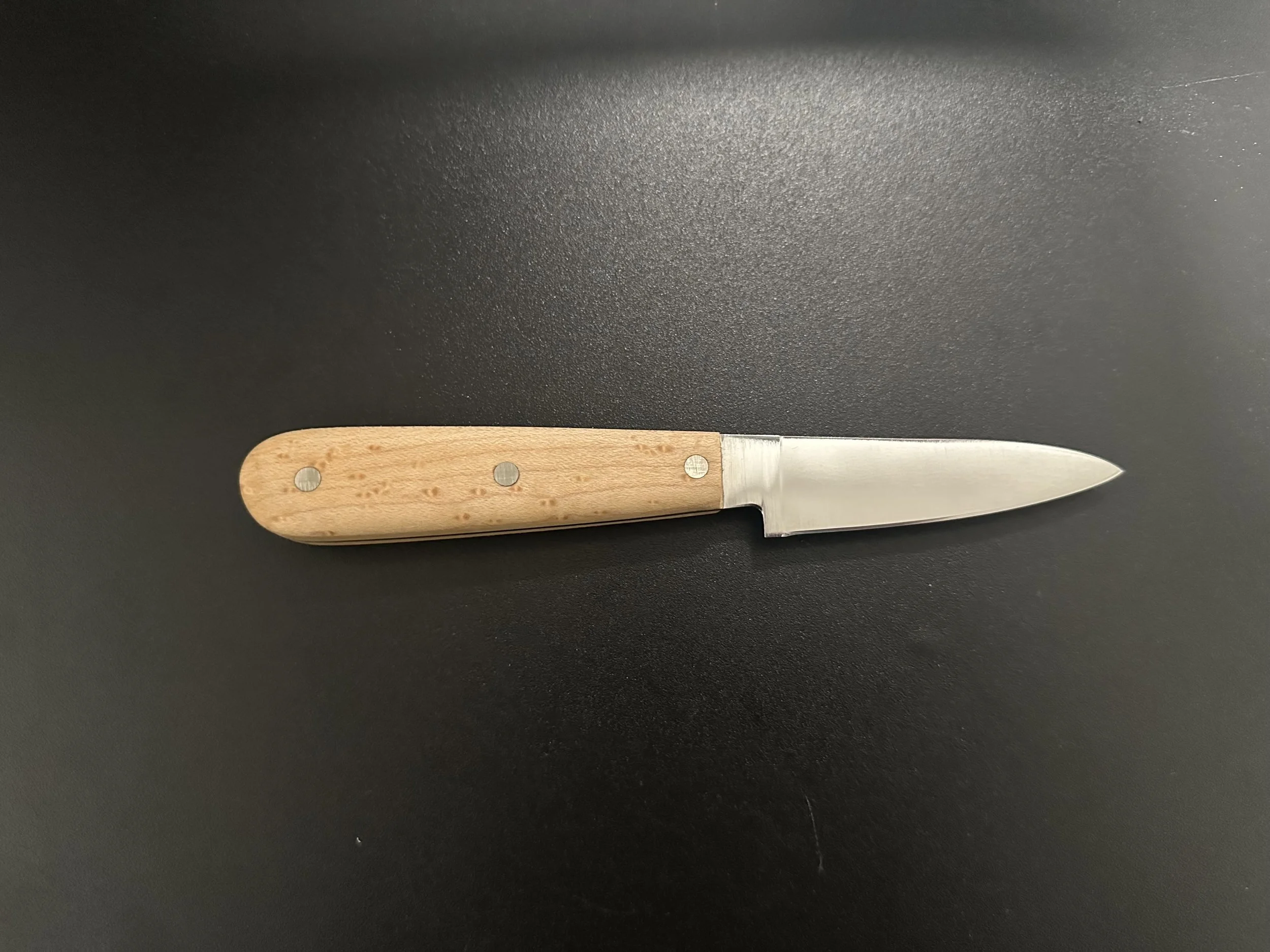 A small knife with a light wooden handle and a stainless steel blade, lying on a black surface.