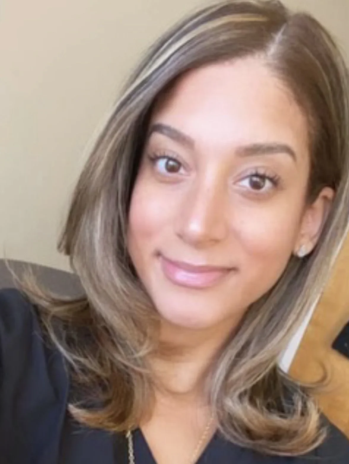 A woman with shoulder-length brown hair with blonde highlights, smiling, wearing earrings and a black top.