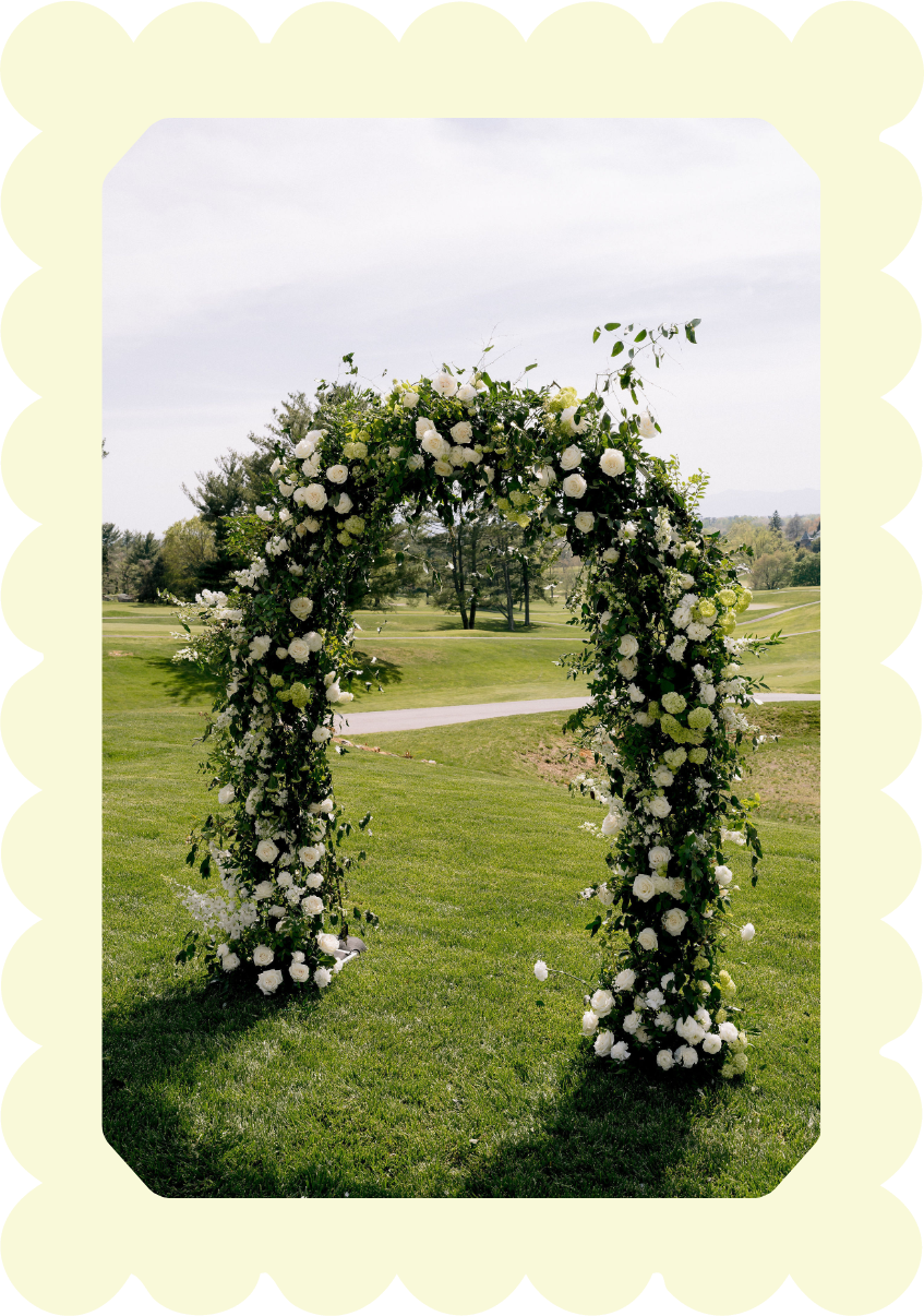 Flower arch decorated with white roses and greenery on a grassy field at a wedding venue.