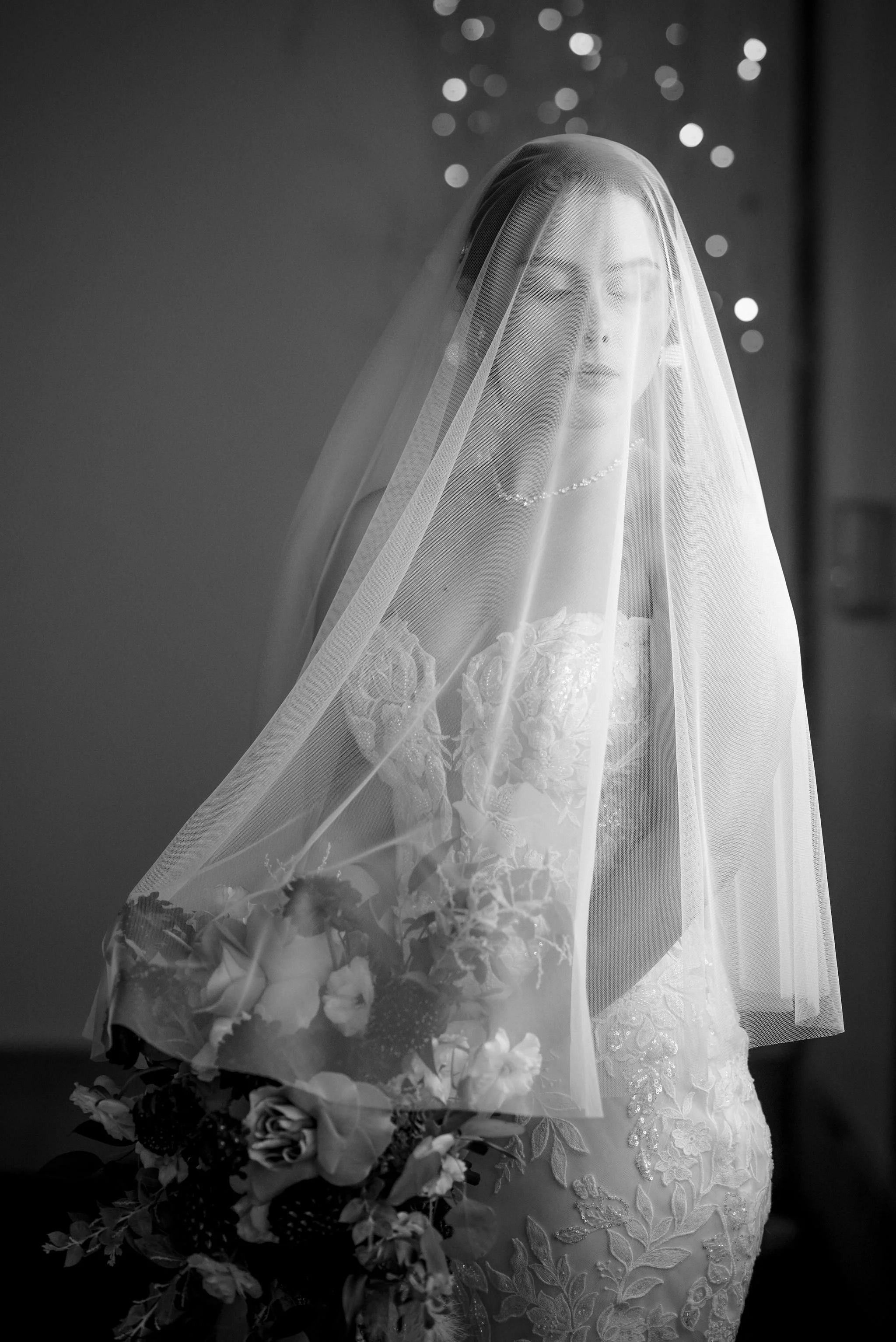 Black and white photo of a bride with a veil over her face, wearing a lace wedding dress, holding a bouquet, with soft lighting and bokeh lights in the background.