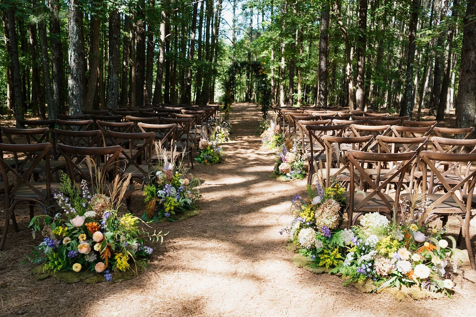 Outdoor wedding ceremony setup in a forest with flower arrangements along the aisle, chairs on either side, and an arch in the background.