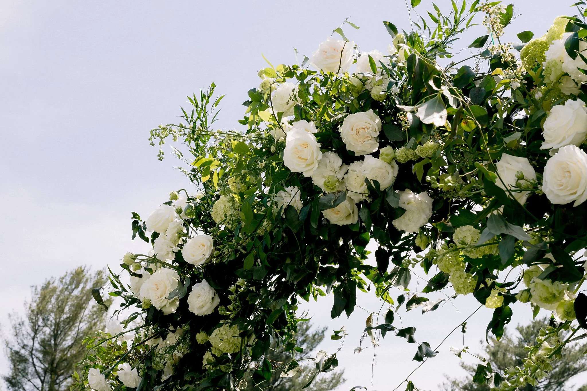 A floral arrangement with white roses, white hydrangeas, and greenery hanging outdoors against a cloudy sky.
