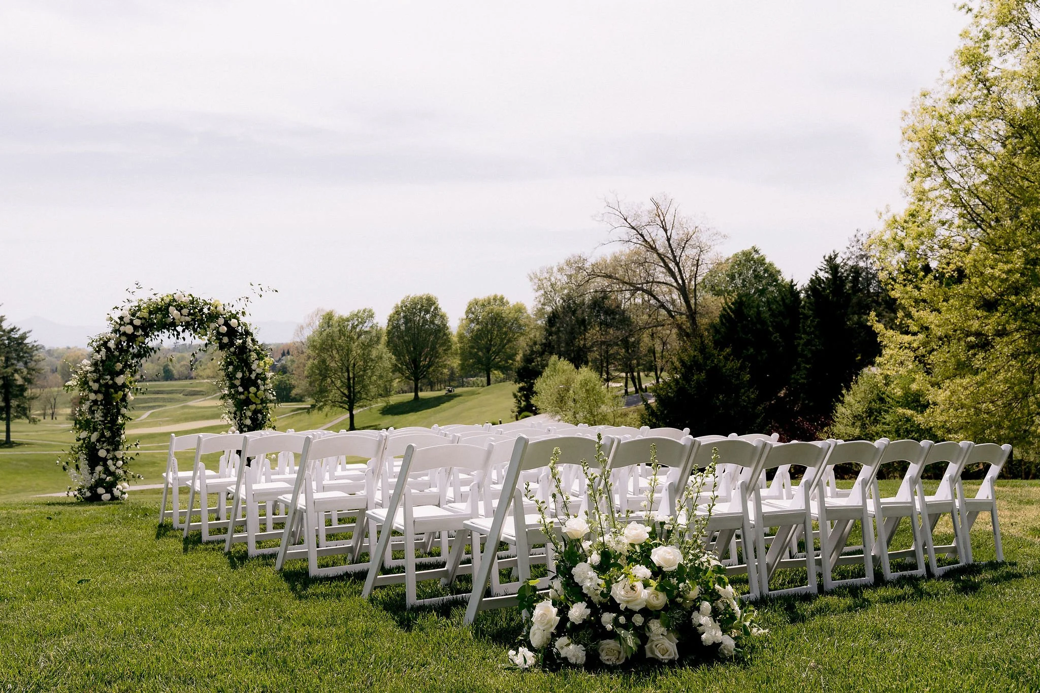 An outdoor wedding setup on a grassy field with white chairs arranged in rows, a floral arch, and a bouquet of white roses in front, with trees and a golf course in the background on a cloudy day.