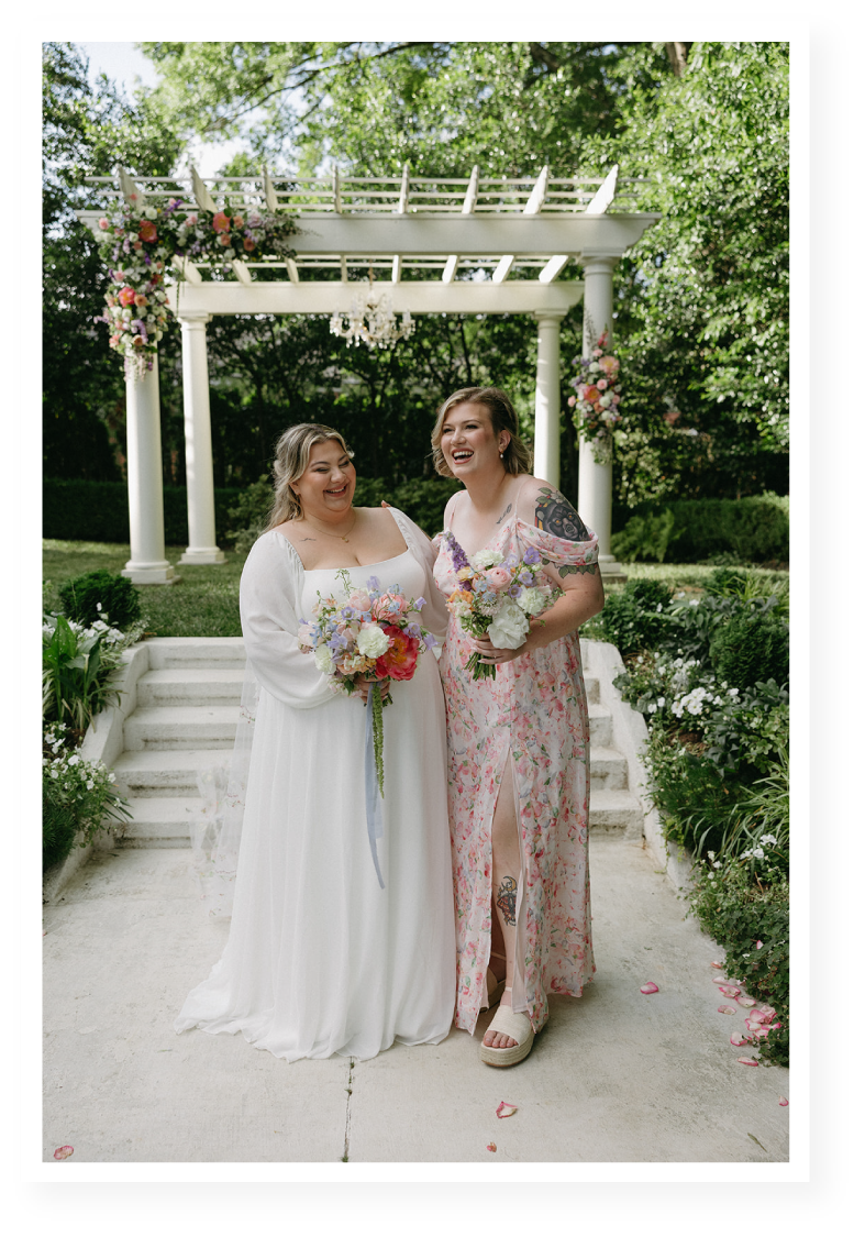 Two women in wedding attire holding bouquets, standing under a floral decorated arch in a garden setting, laughing and celebrating.