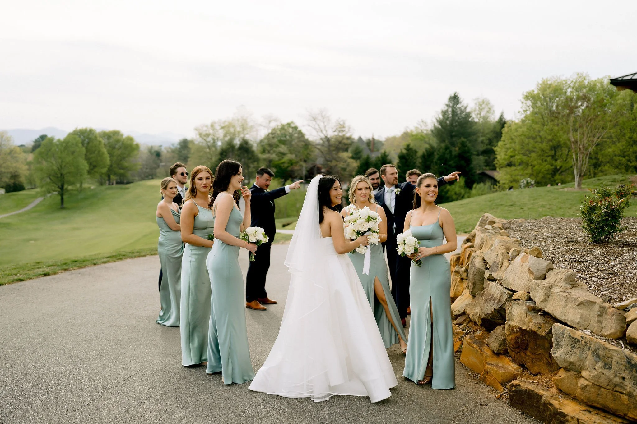 A bride and wedding party standing outdoors on a paved path, with the bride in a white gown and veil and the bridesmaids in matching light blue dresses holding white bouquets. The groom and groomsmen are dressed in dark suits, some pointing and smili