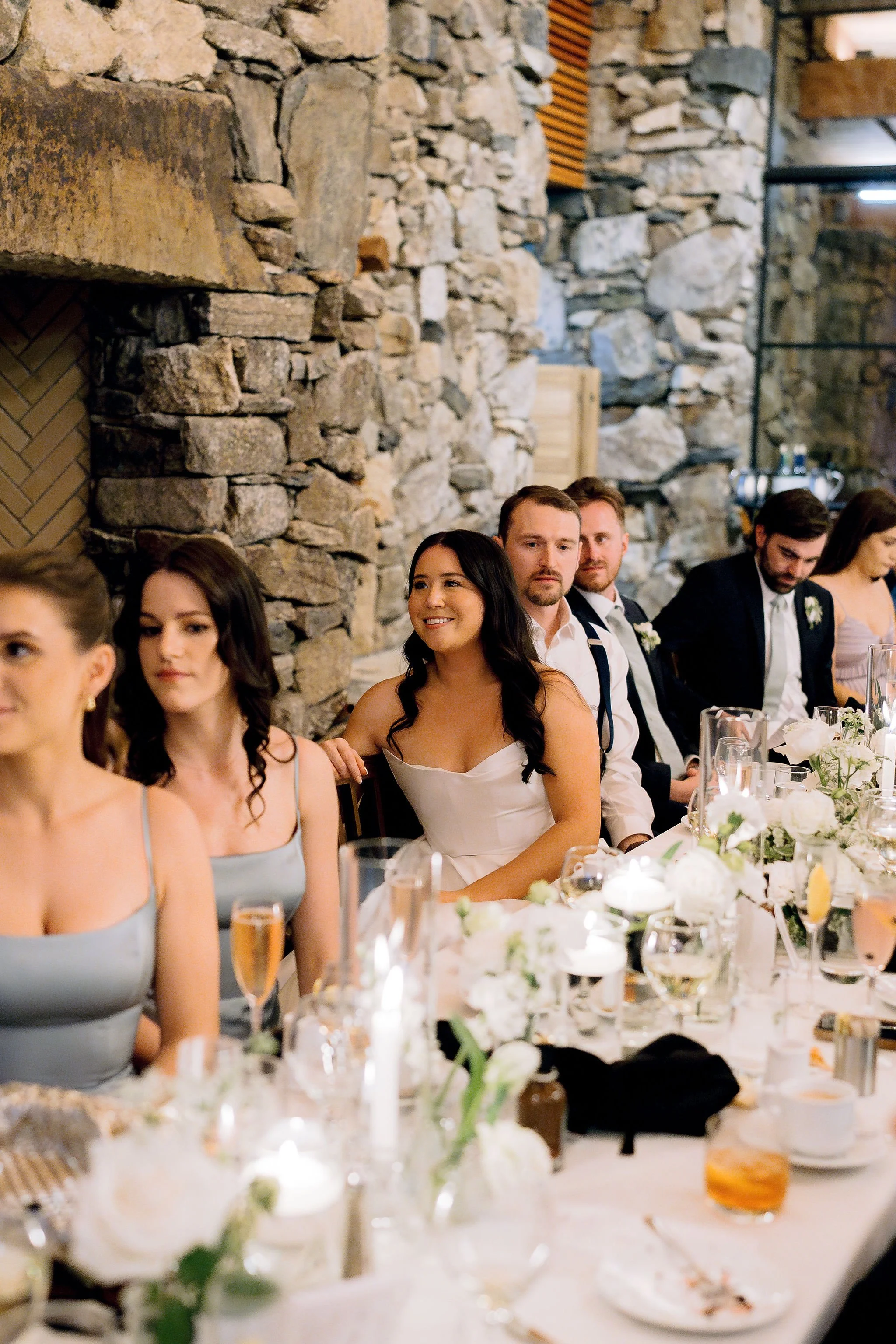 People sitting at a long table during a wedding reception, with a stone wall in the background, decorated with white flowers and candles.