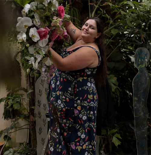 A woman with long dark hair in a floral dress arranging pink and white flowers in a lush indoor garden.