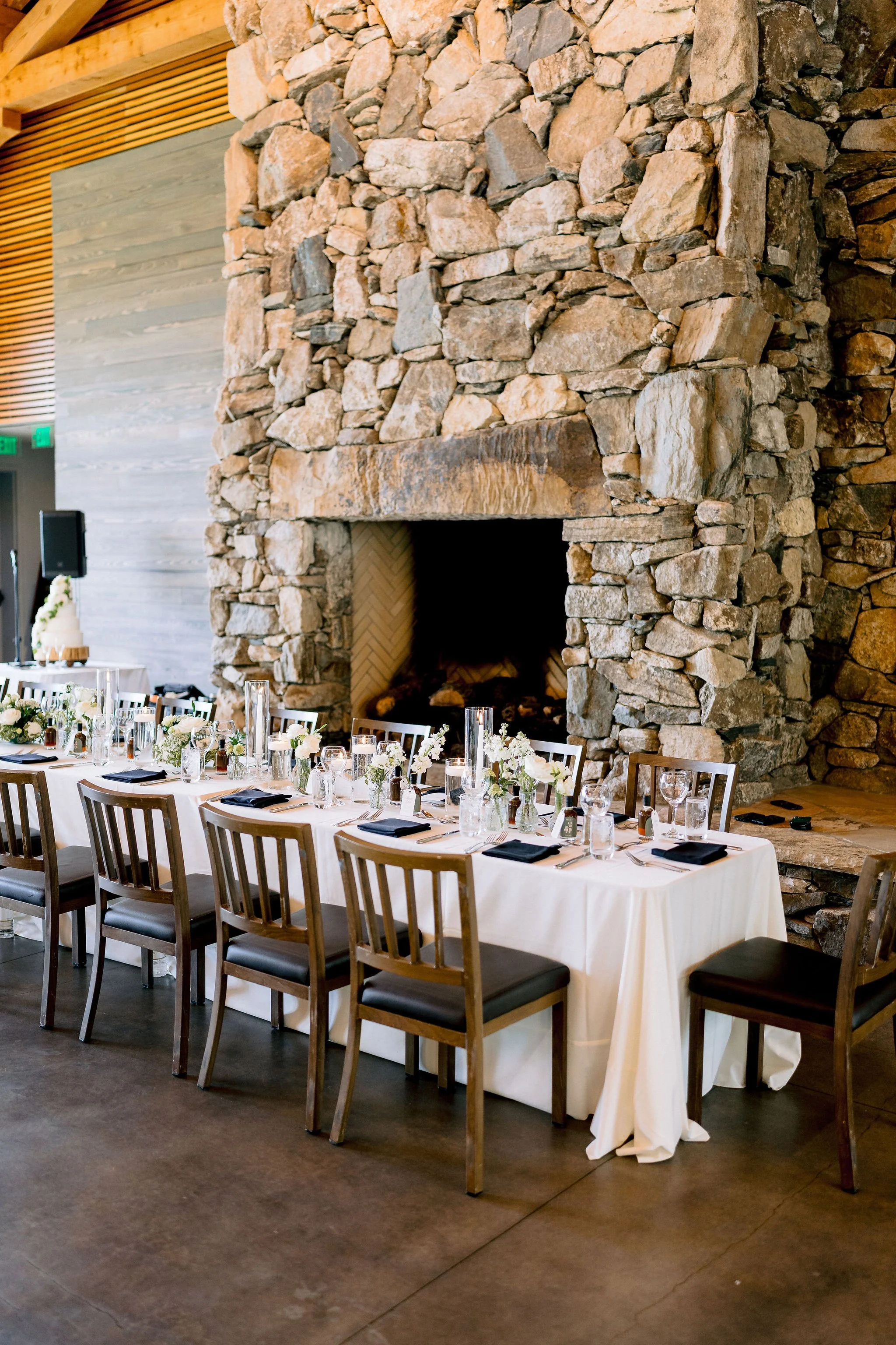A long banquet table set for a meal in front of a large stone fireplace in a rustic venue, with white tablecloths, black napkins, glassware, and floral centerpieces.