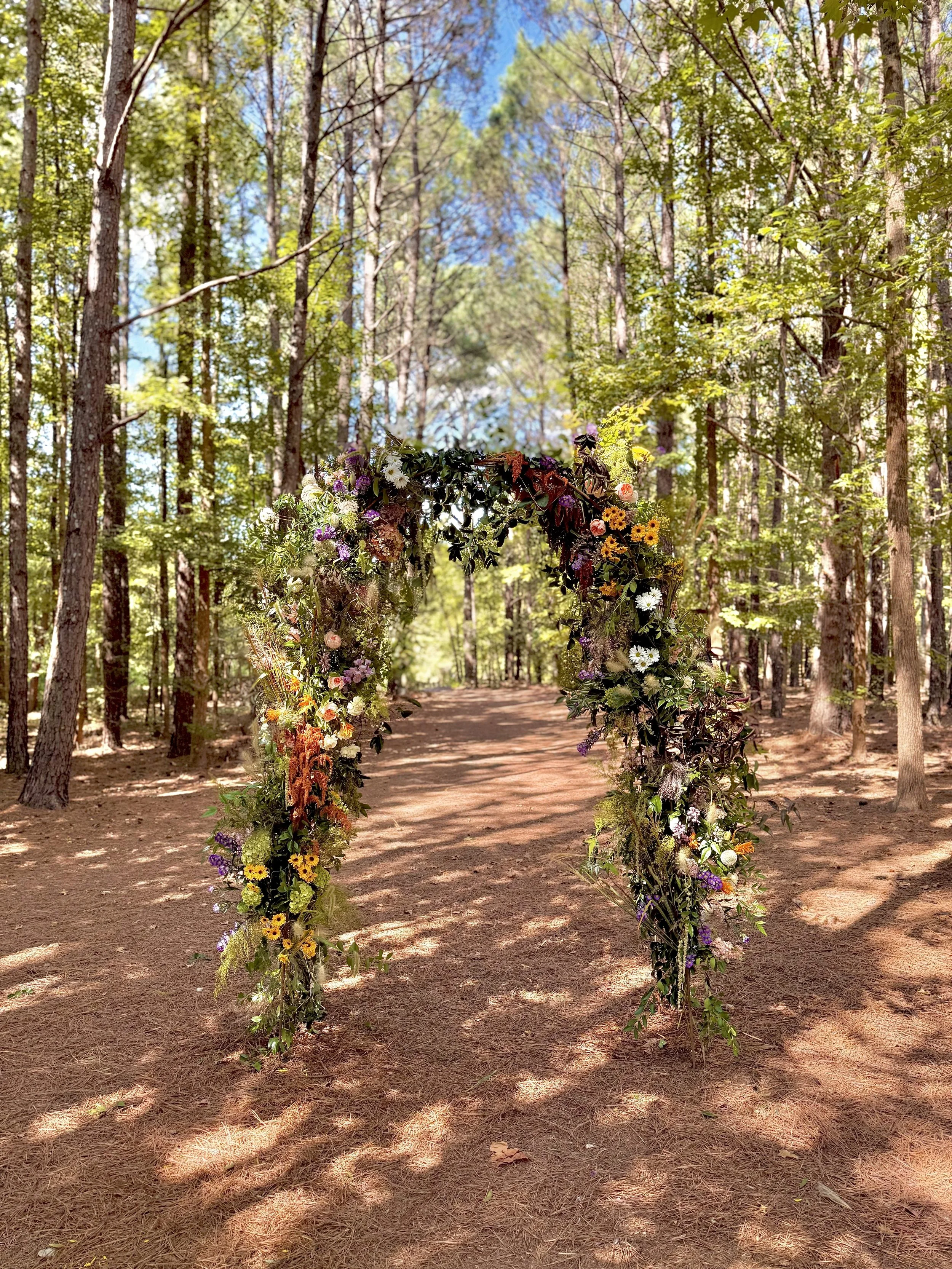 A floral arch placed on a dirt pathway in a forest, surrounded by tall trees and green foliage, with sunlight filtering through the leaves.