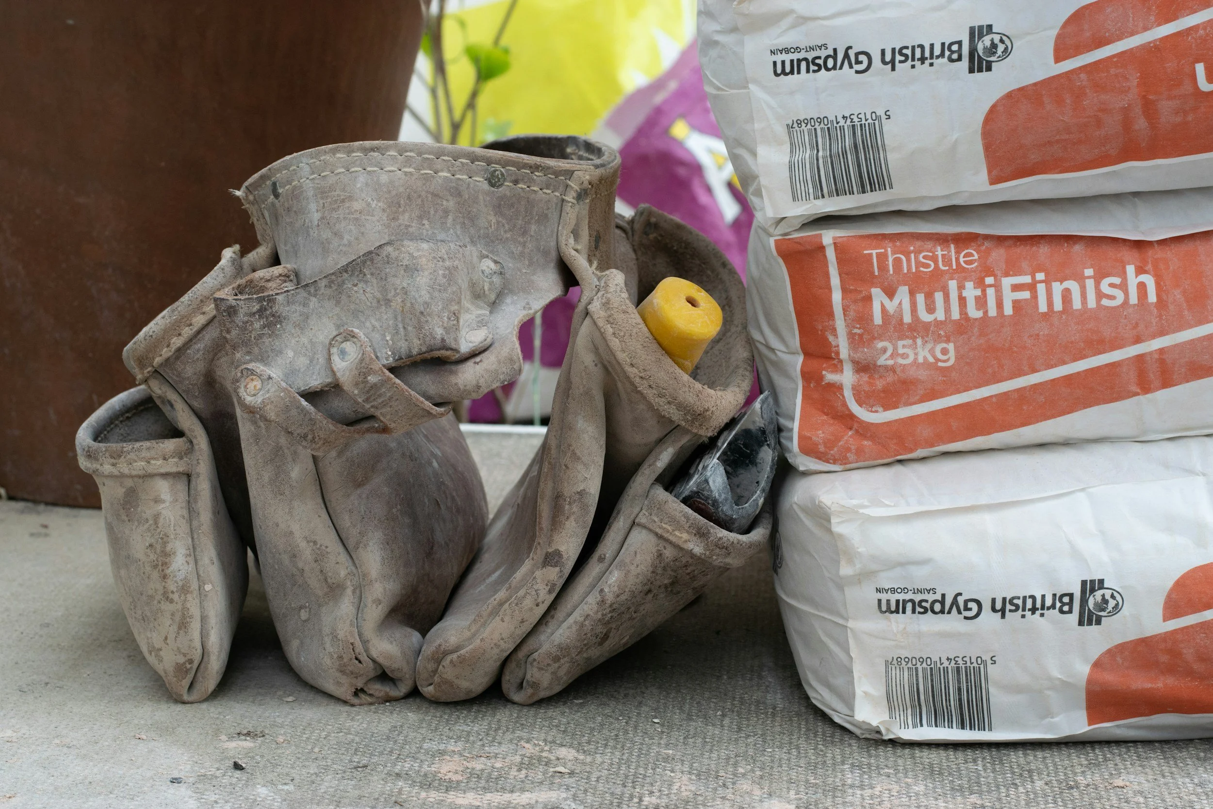 A pair of well-worn, dusty baseball gloves stacked next to a bag of Thistle MultiFinish, 25 kg, on a concrete surface.