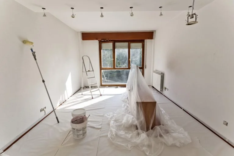 Empty room undergoing renovation, with a step ladder, window, paint roller, bucket, and a piece of furniture covered with plastic sheet.