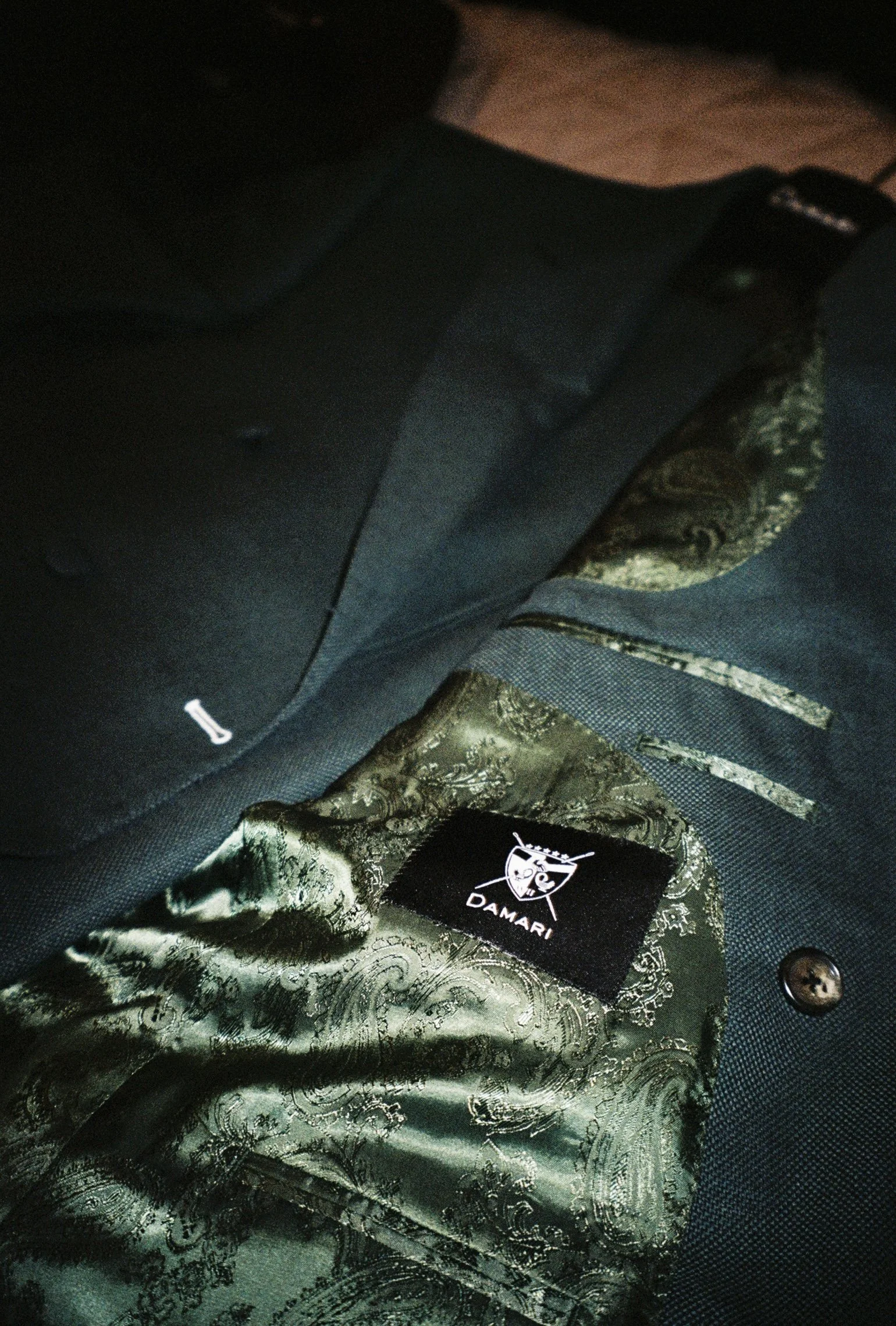 Close-up of a textile label on a black fabric jacket, showing the brand name 'DAMARI' and a logo of a shield with a sword, along with ornate patterned lining inside the jacket.