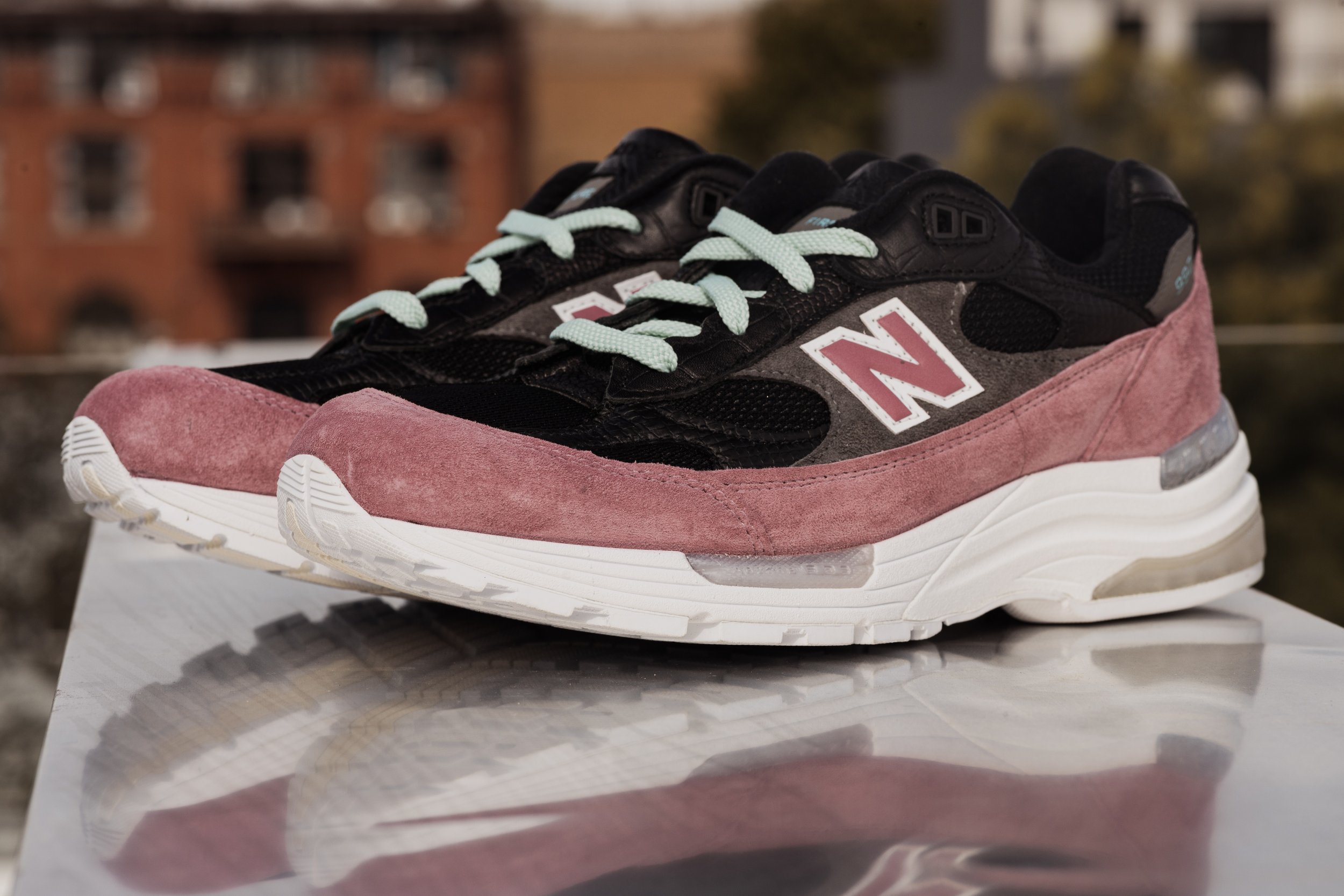 A pair of New Balance sneakers with black, pink, and grey tones, featuring mint green laces, placed outdoors on a reflective surface with buildings and trees in the background.