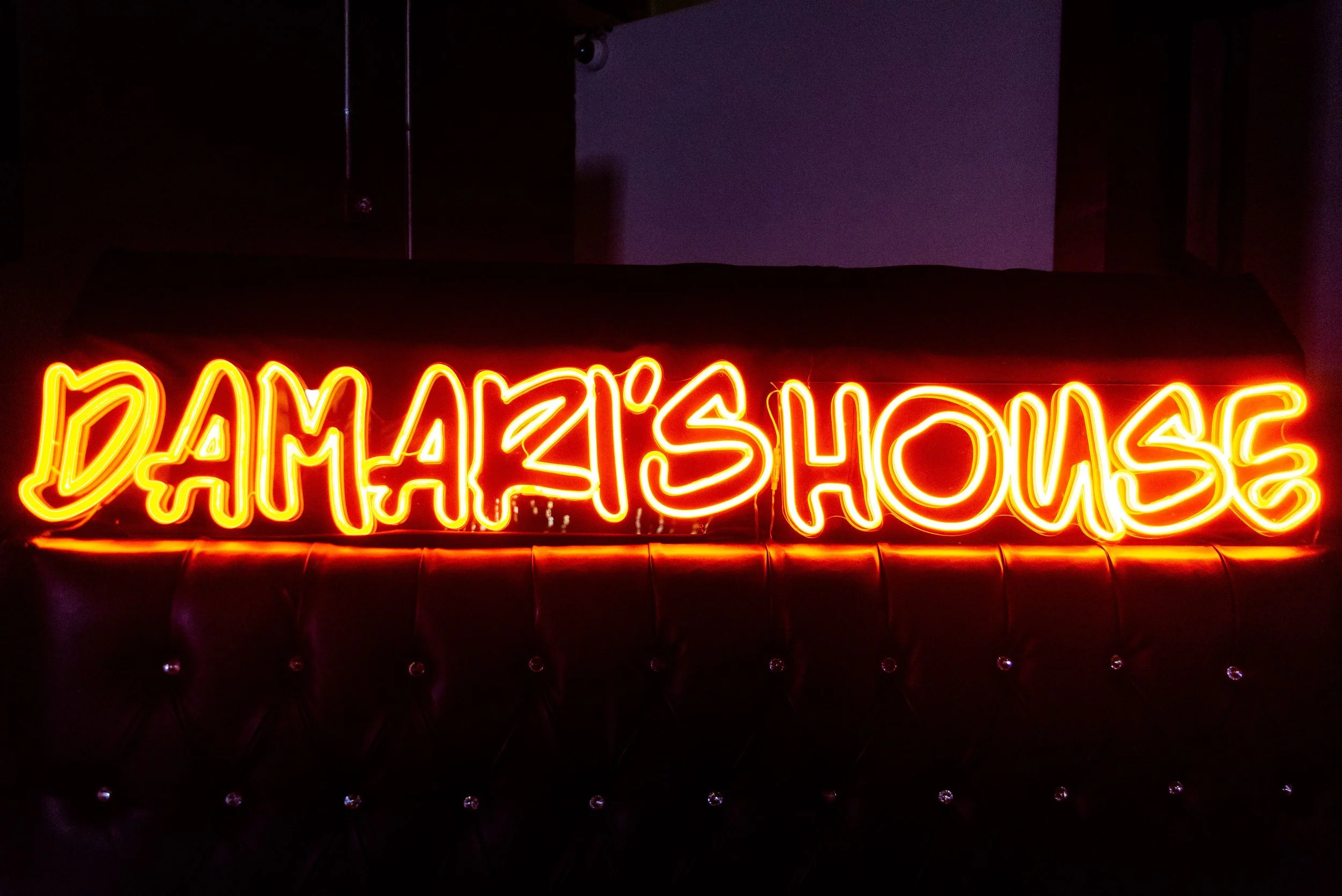 Neon sign in bright yellow and red colors displaying the words 'DAMA'S HOUSE' on a dark background.
