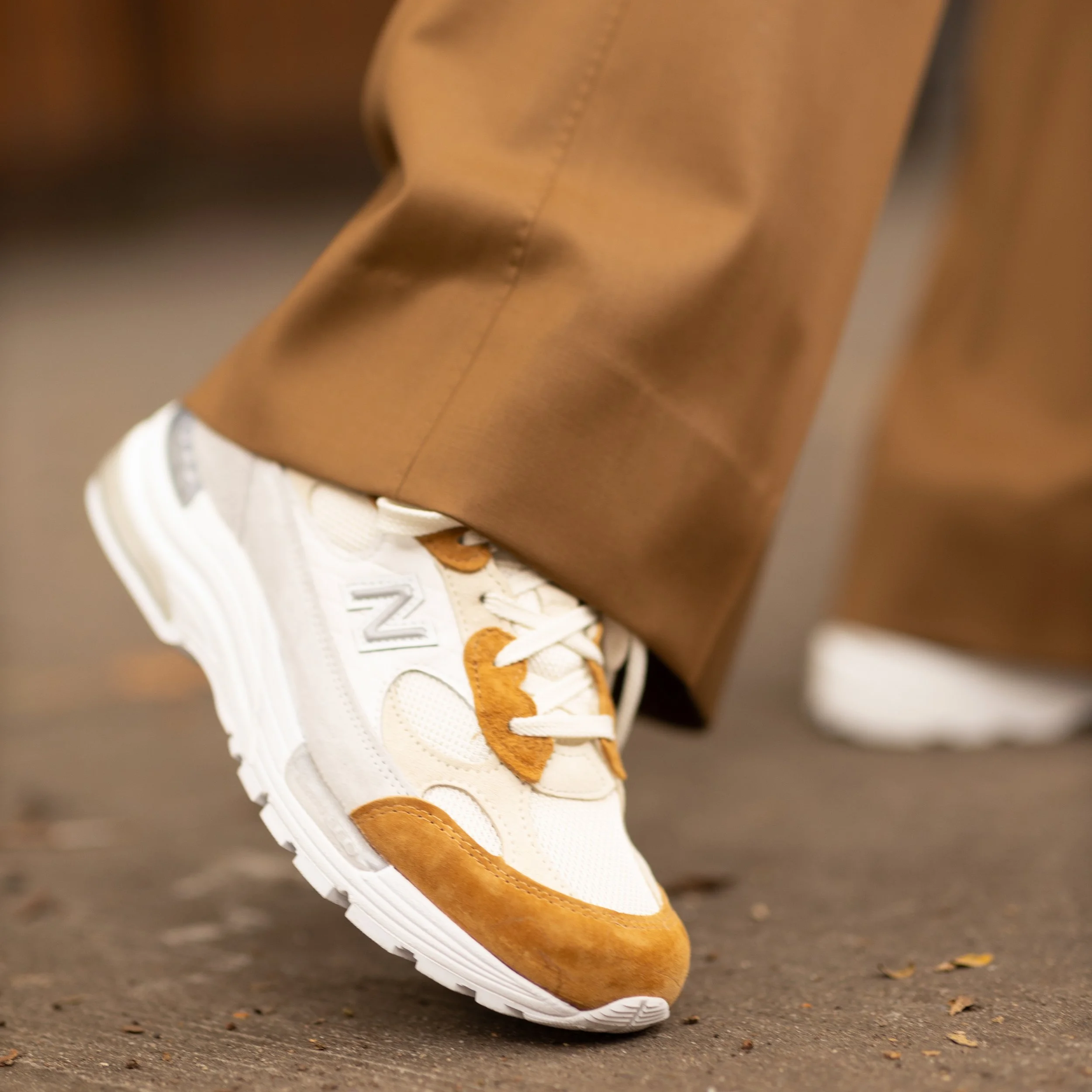 Close-up of a white sneaker with tan accents and brown suede detailing, worn with brown pants.