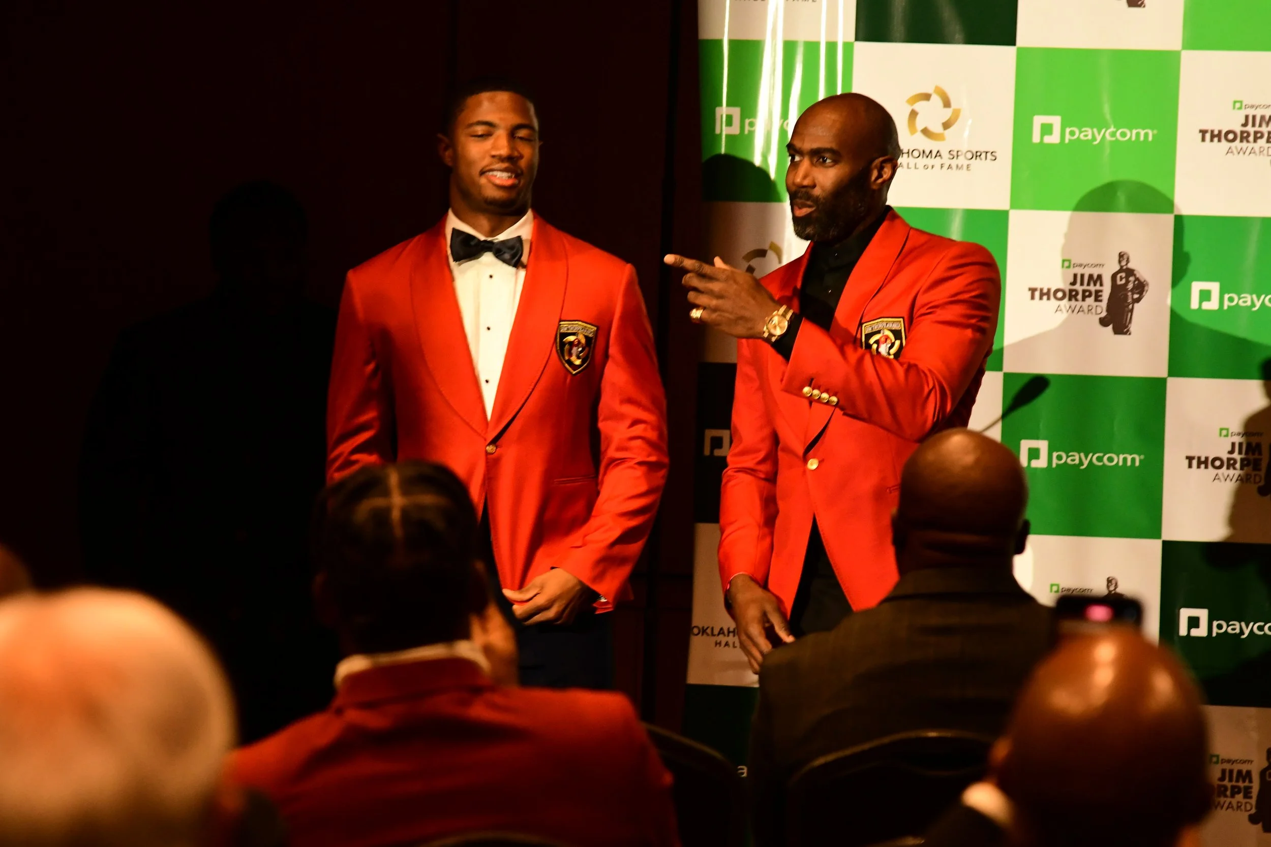 Two men dressed in red tuxedos speaking at an awards event with a backdrop featuring sponsor logos and event branding.