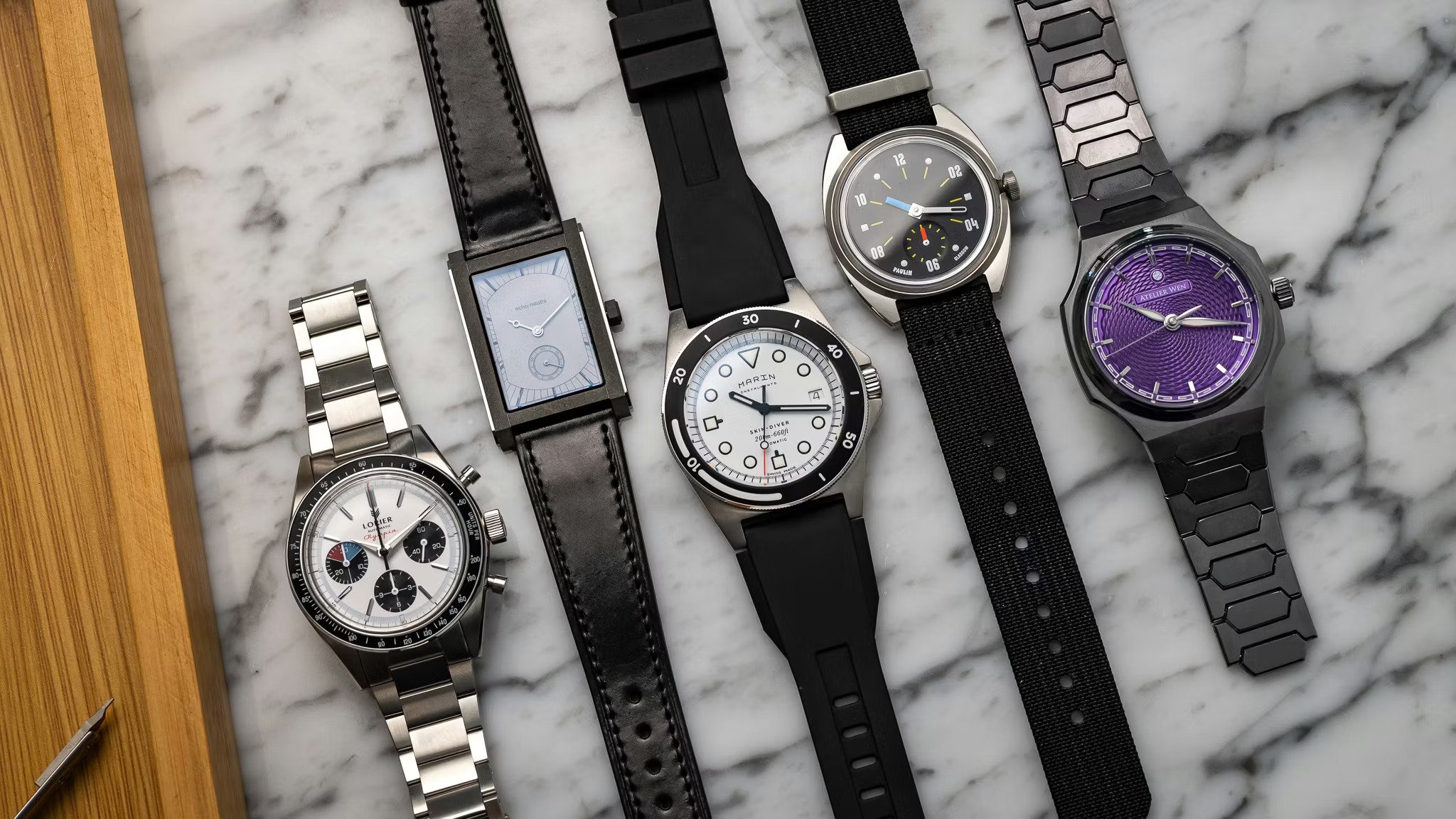 Five independent watches on a marble surface, including a Lorier chronograph, rectangular dress watch, Martenero diver, Serica field watch, and purple Atelier Wen