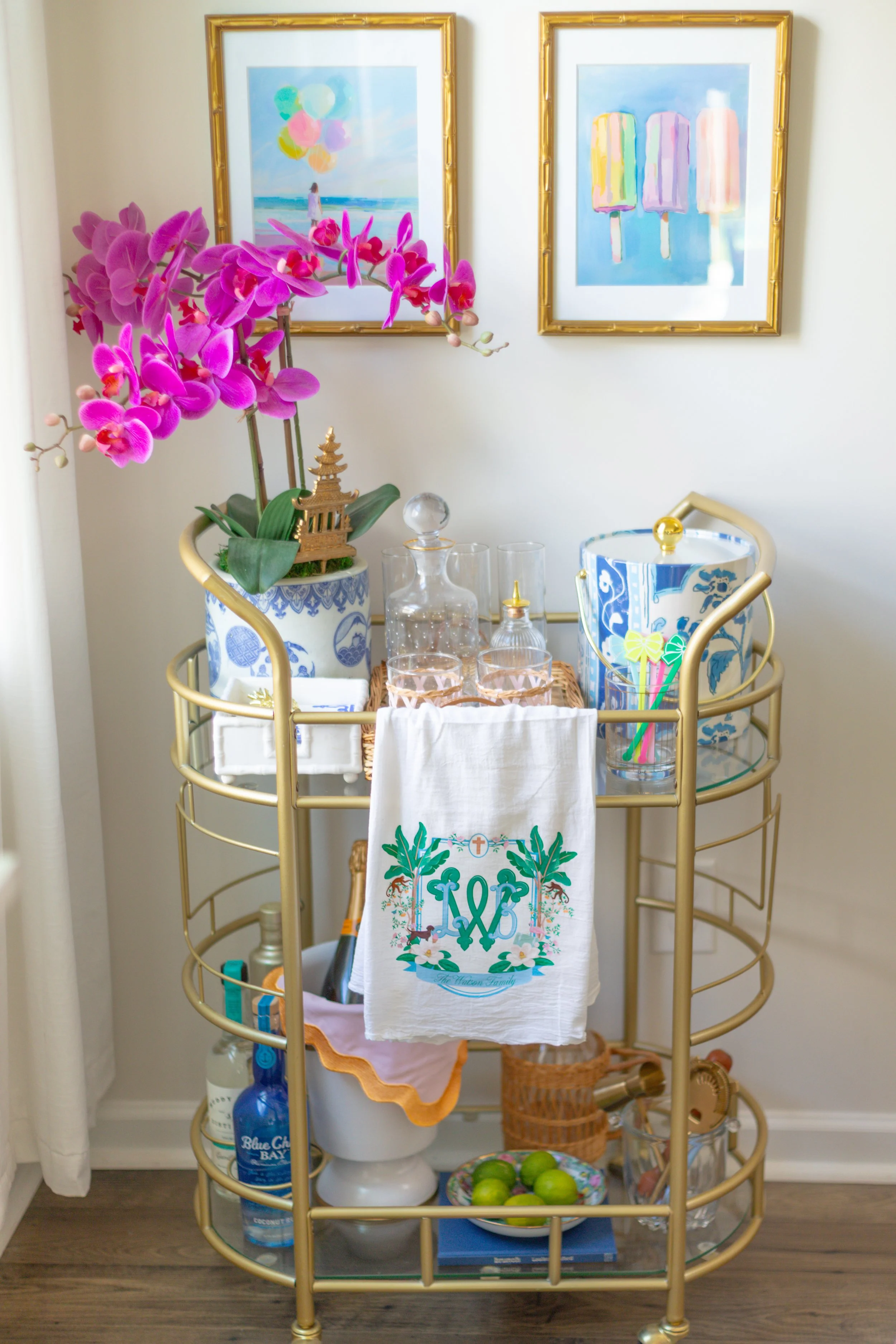 A gold bar cart with various glasses, a decanter, and a towel with a monogram, placed against a white wall with two framed beach-themed watercolors, and decorated with a pink orchid plant.