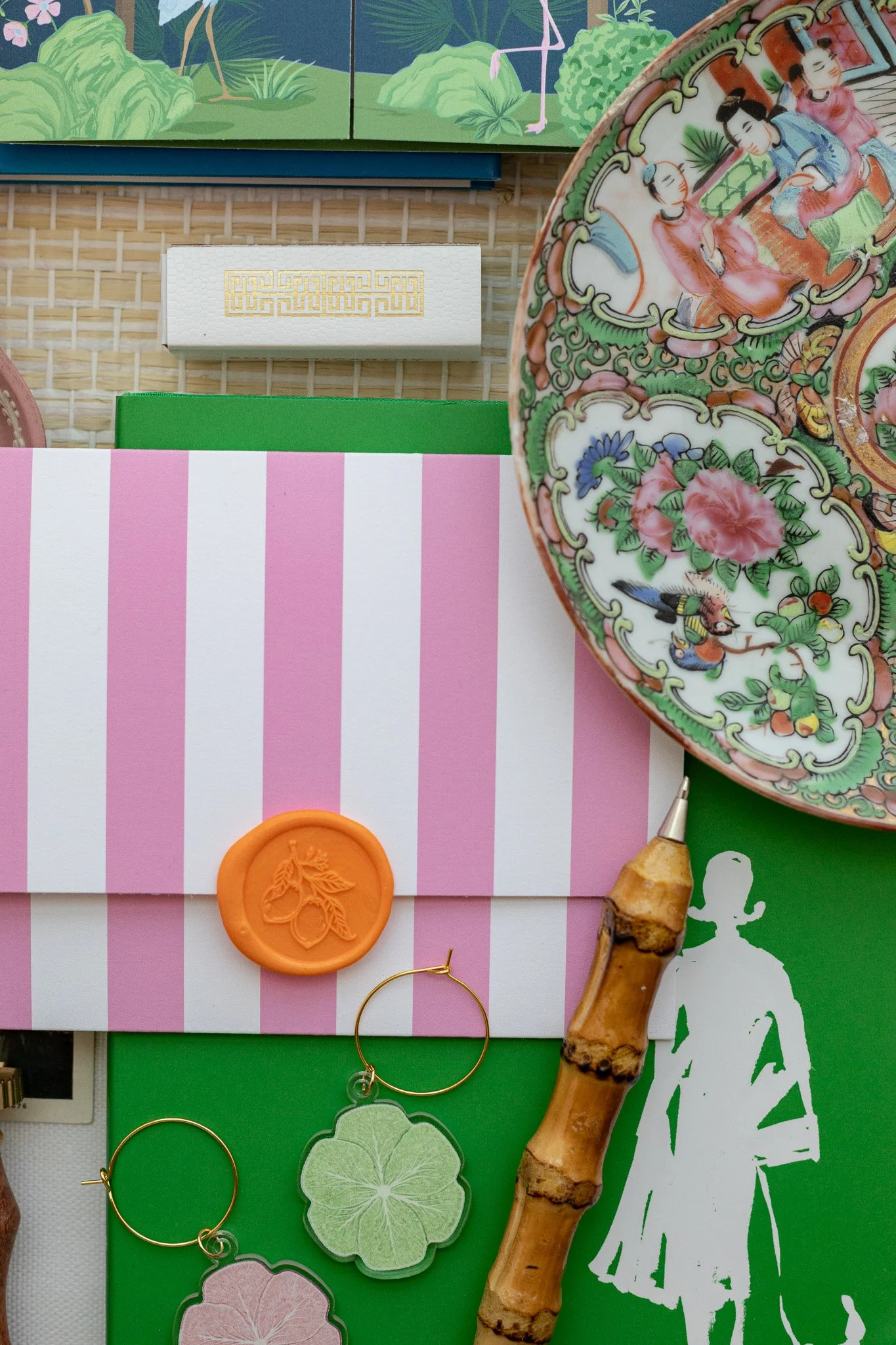 A flat lay of decorative items including a colorful Asian porcelain plate, pink and white striped box with a wax seal, gold hoop earrings, a green leafy cutout, a bamboo stick, and a green book, with a storage board in the background.