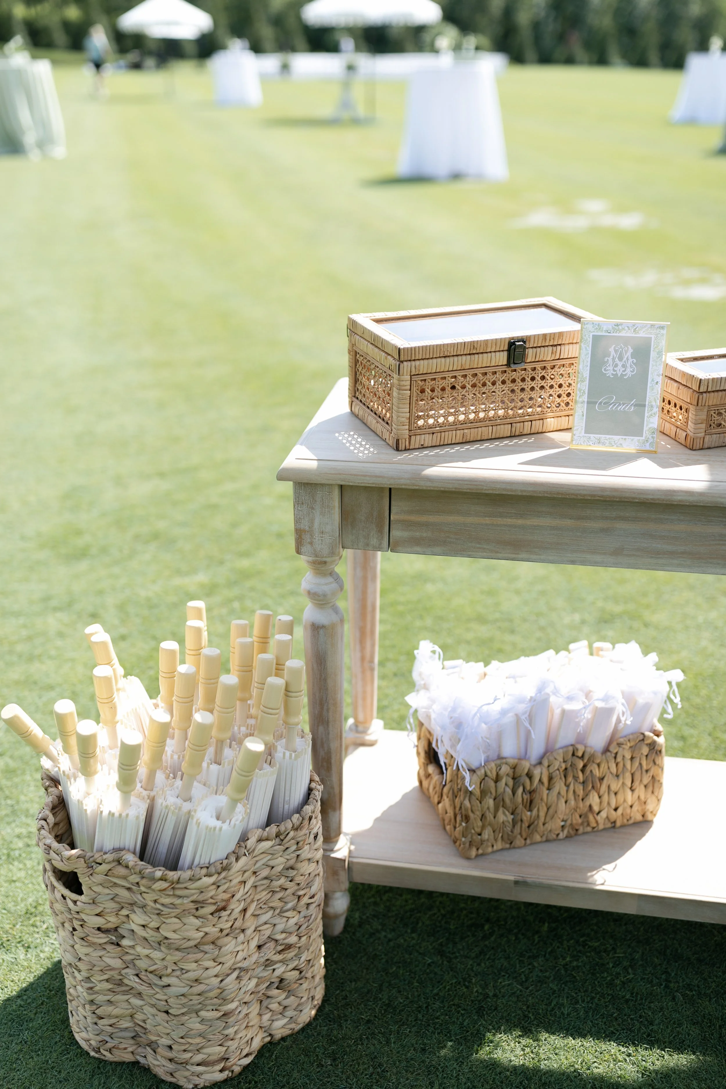 Beach wedding card and tube station decorated with woven baskets of white programs and pens, tables with card signs, on a grassy outdoor setting.