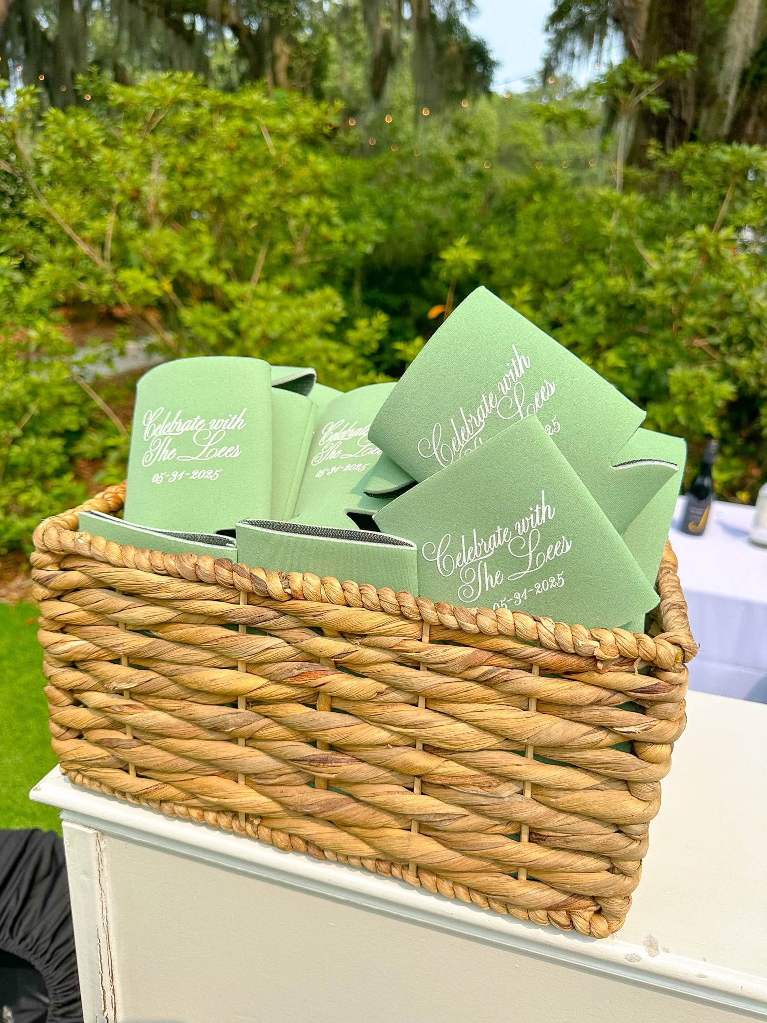 Green wedding programs in a woven basket at an outdoor celebration, with a background of greenery and string lights.