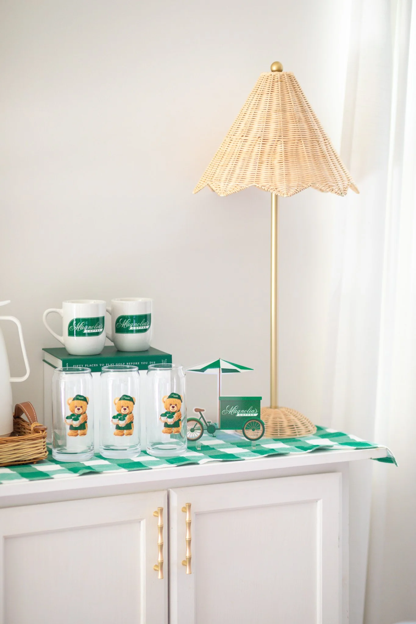 Decorative tabletop display with magnolia-themed items, including two coffee mugs, three glasses with teddy bear images, a small paper food cart with an umbrella, a bicycle, a lamp with a wicker shade, and a plaid tablecloth, all on a white cabinet.