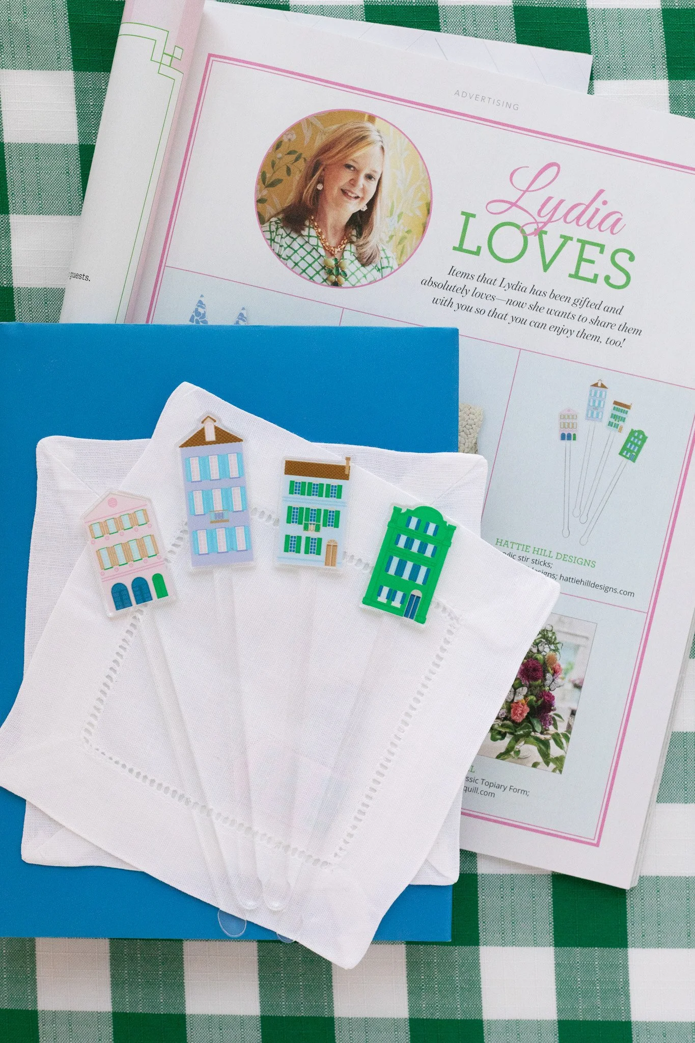 A collection of items including a pamphlet titled "Lydia LOVES" featuring a woman's photo, four colorful miniature building stickers on white napkins, and a blue folder, arranged on a green and white checkered tablecloth.