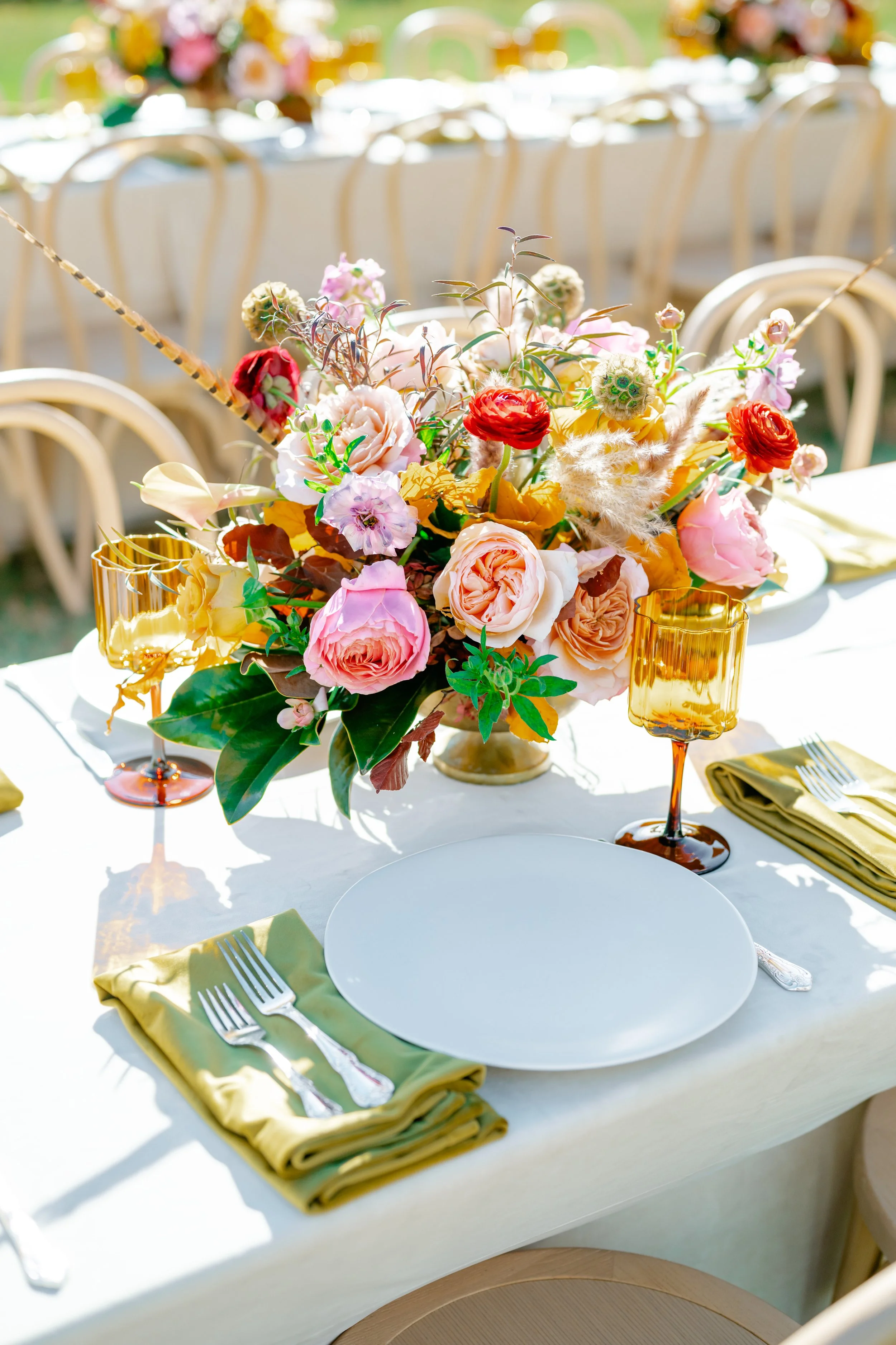 Table decorated with a colorful flower arrangement, gold-rimmed glasses, silverware, and folded yellow napkins at a wedding or event.
