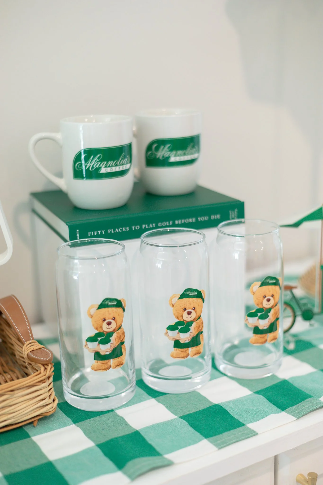 Decorative display with two white coffee mugs featuring green logos, three clear glasses with bear illustrations, a green book titled 'Fifty Places to Play Golf Before You Die,' and a small wooden toy bicycle on a green and white checkered tablecloth
