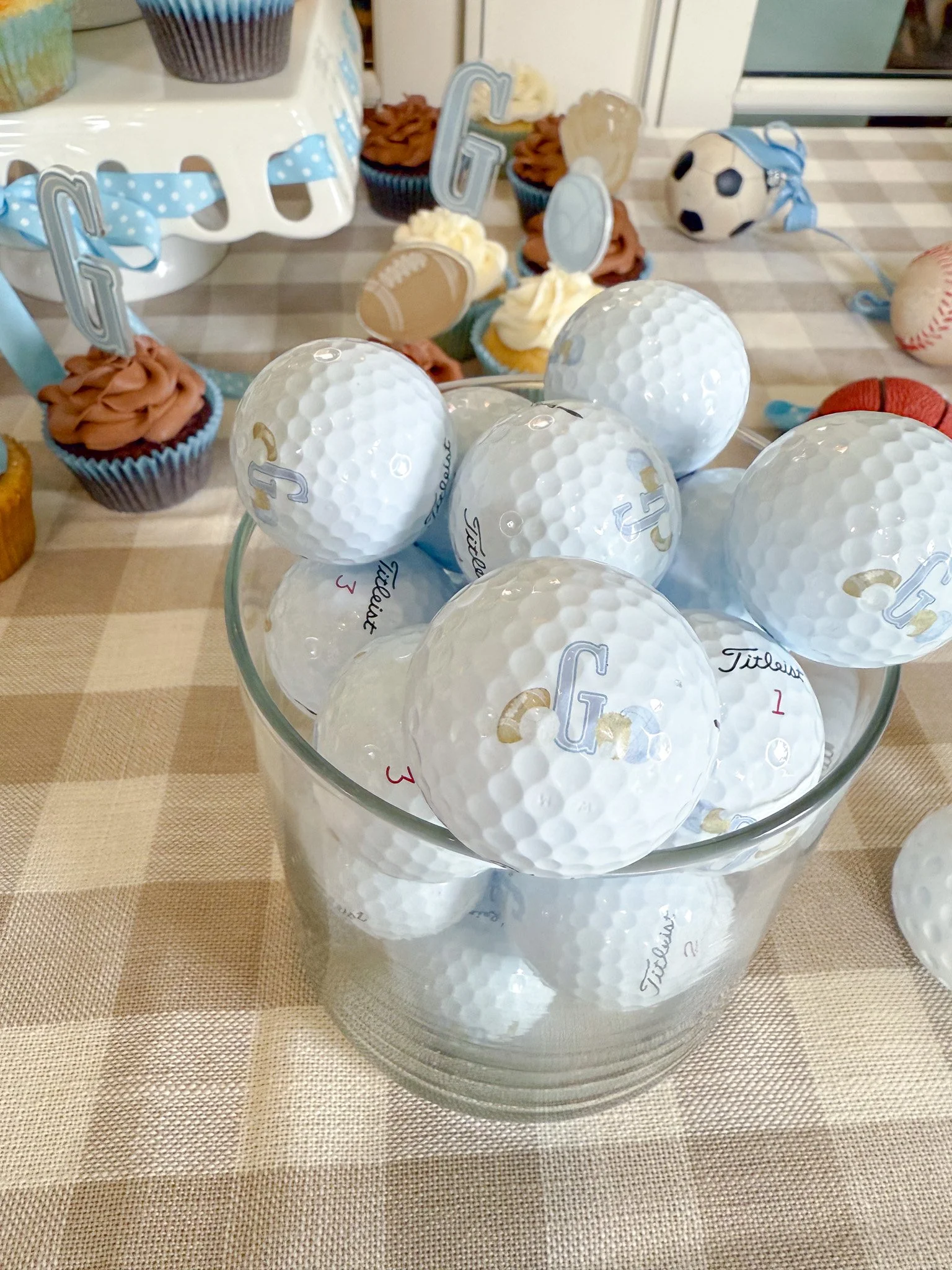 A glass bowl filled with white golf balls, with some inside and some on top. In the background, a dessert table with cupcakes decorated with sports-themed toppers, and various small sports-themed decorations such as a soccer ball and a baseball.