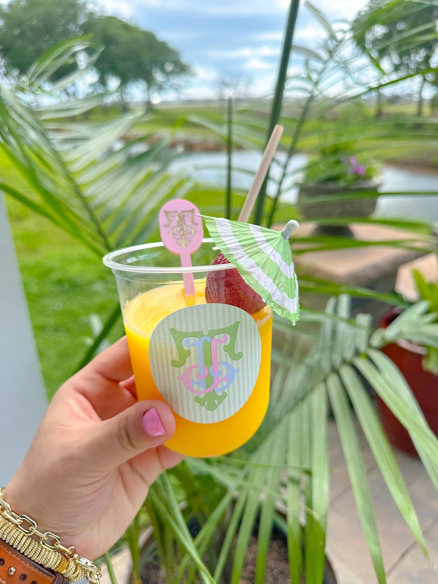 A hand holding a clear plastic cup with yellow liquid, a strawberry, a green and white paper umbrella, and a straw, with a colorful decorative label. In the background, there is green foliage and a pond or water feature outdoors.