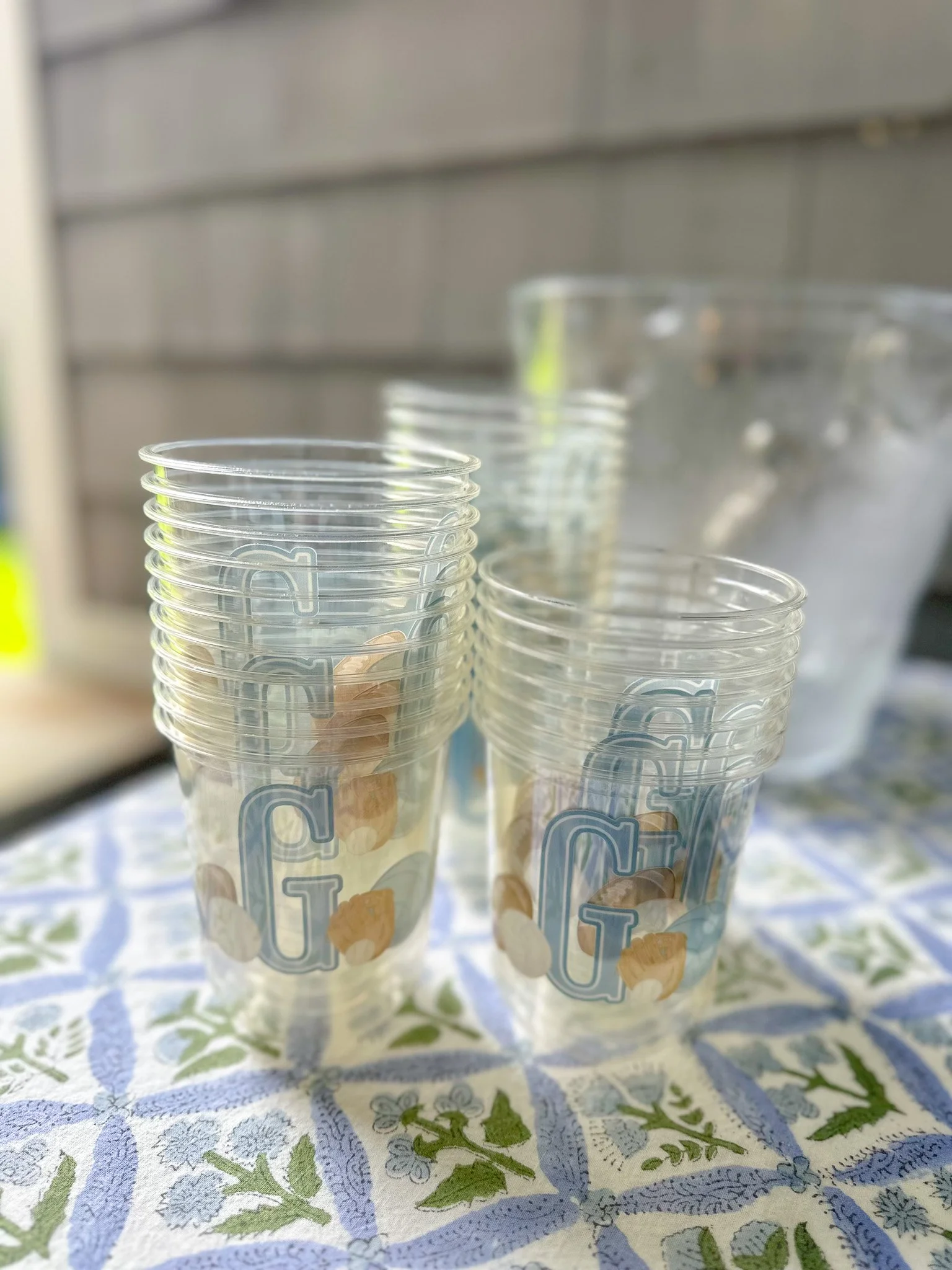 Stacked plastic cups with a glass pitcher in the background on a table with a blue and green patterned tablecloth.