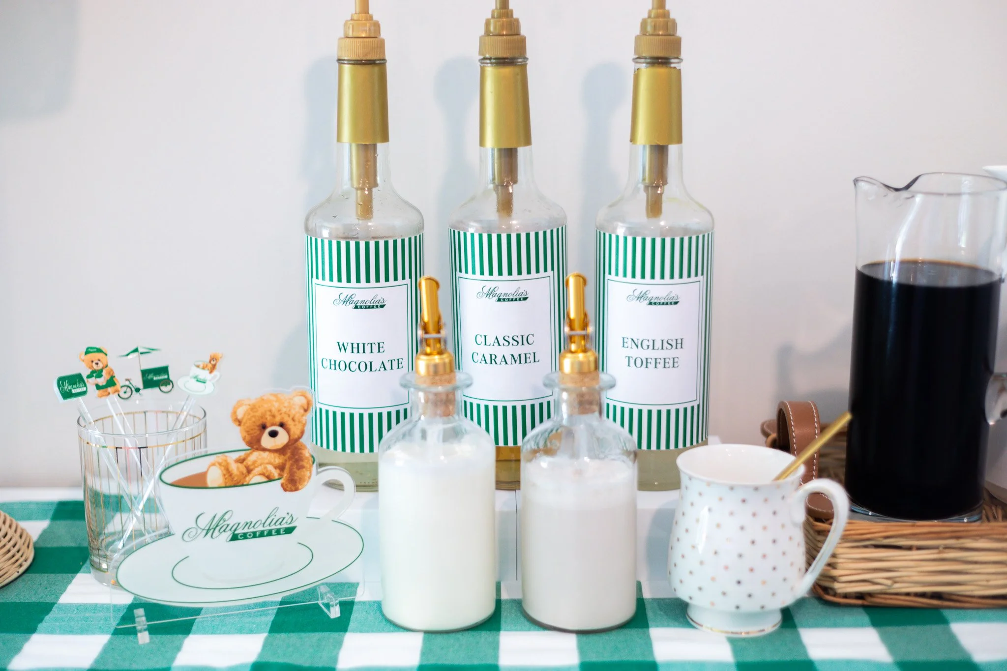 Assorted coffee syrups labeled 'White Chocolate,' 'Classic Caramel,' and 'English Toffee' on a table with a checkered green and white tablecloth, featuring a mug with a teddy bear and drink stirrer, a small glass with wooden stirrers, a pitcher of co