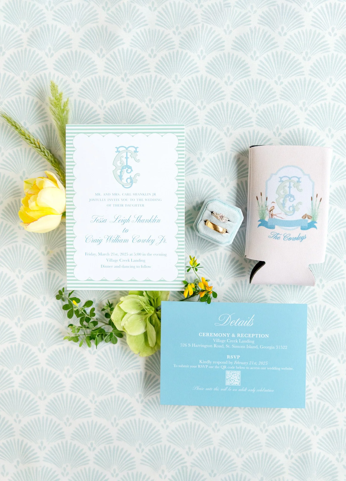 Wedding invitation with floral decorations, rings in a ring box, a cake with a monogram, and an RSVP card on a patterned tablecloth.