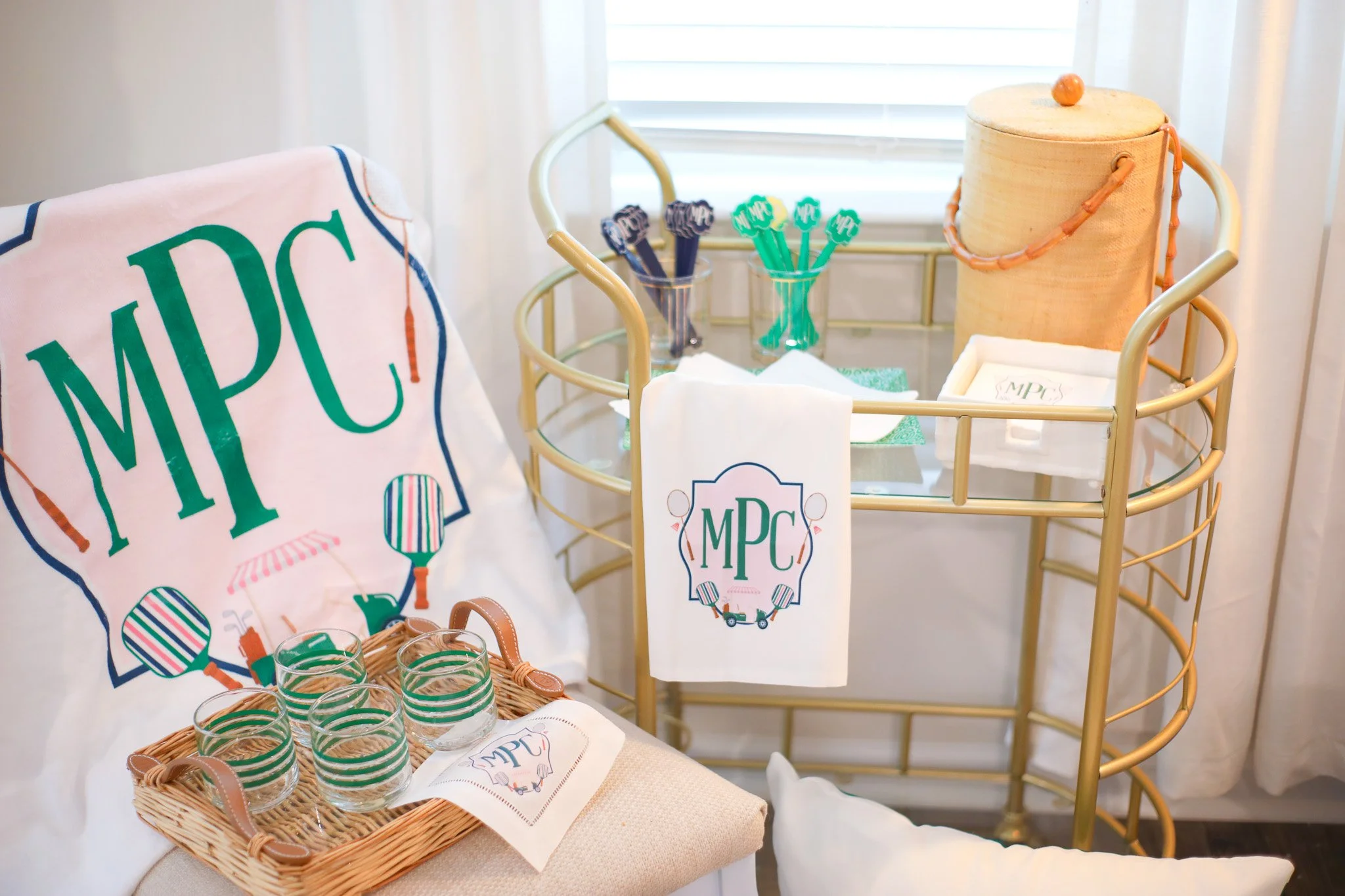 A baby shower setup with a gold cart displaying a beige umbrella-shaped container, green and purple cups, and napkins with the event's logo. Next to the cart, a chair holds a white cloth with a large green and blue monogram "MPC," and a tray of six g