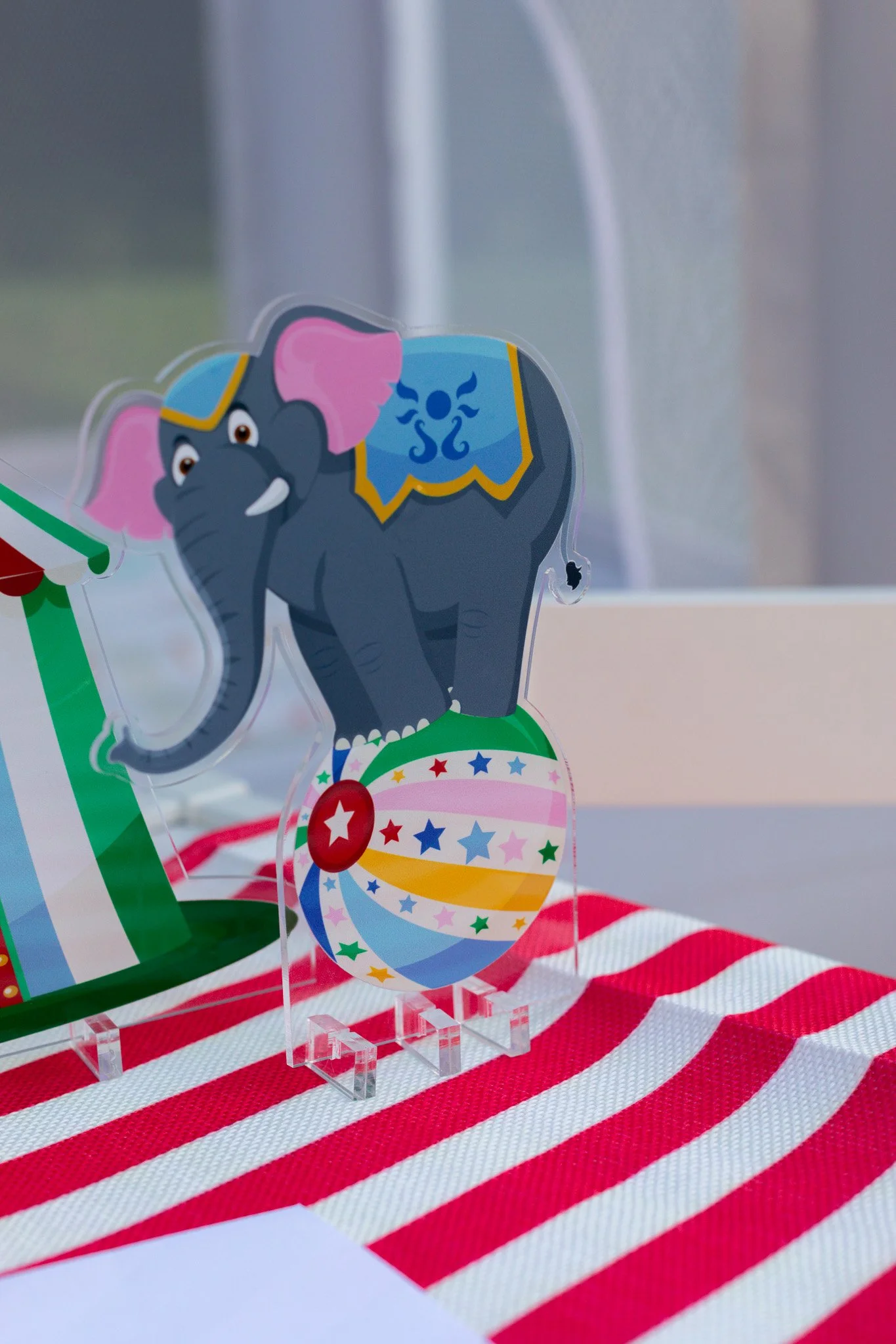 Decorative party box featuring a cartoon elephant standing on a colorful patterned ball, placed on a red and white striped tablecloth.
