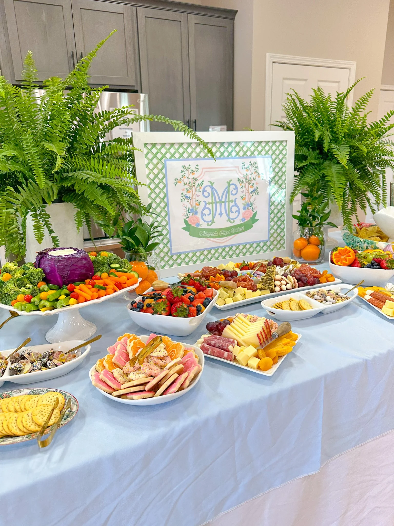 A dessert and fruit table with various sweets, cheeses, fruits, and vegetables, decorated with greenery and framed artwork in an indoor setting.